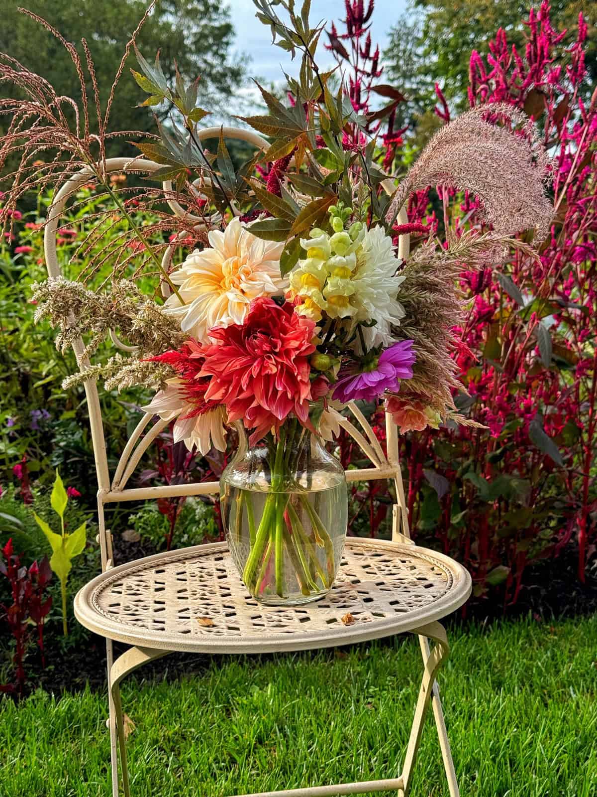 A colorful floral arrangement with red, pink, yellow, and white flowers sits in a glass vase on a vintage metal chair. The background features vibrant greenery and tall, blooming plants.
