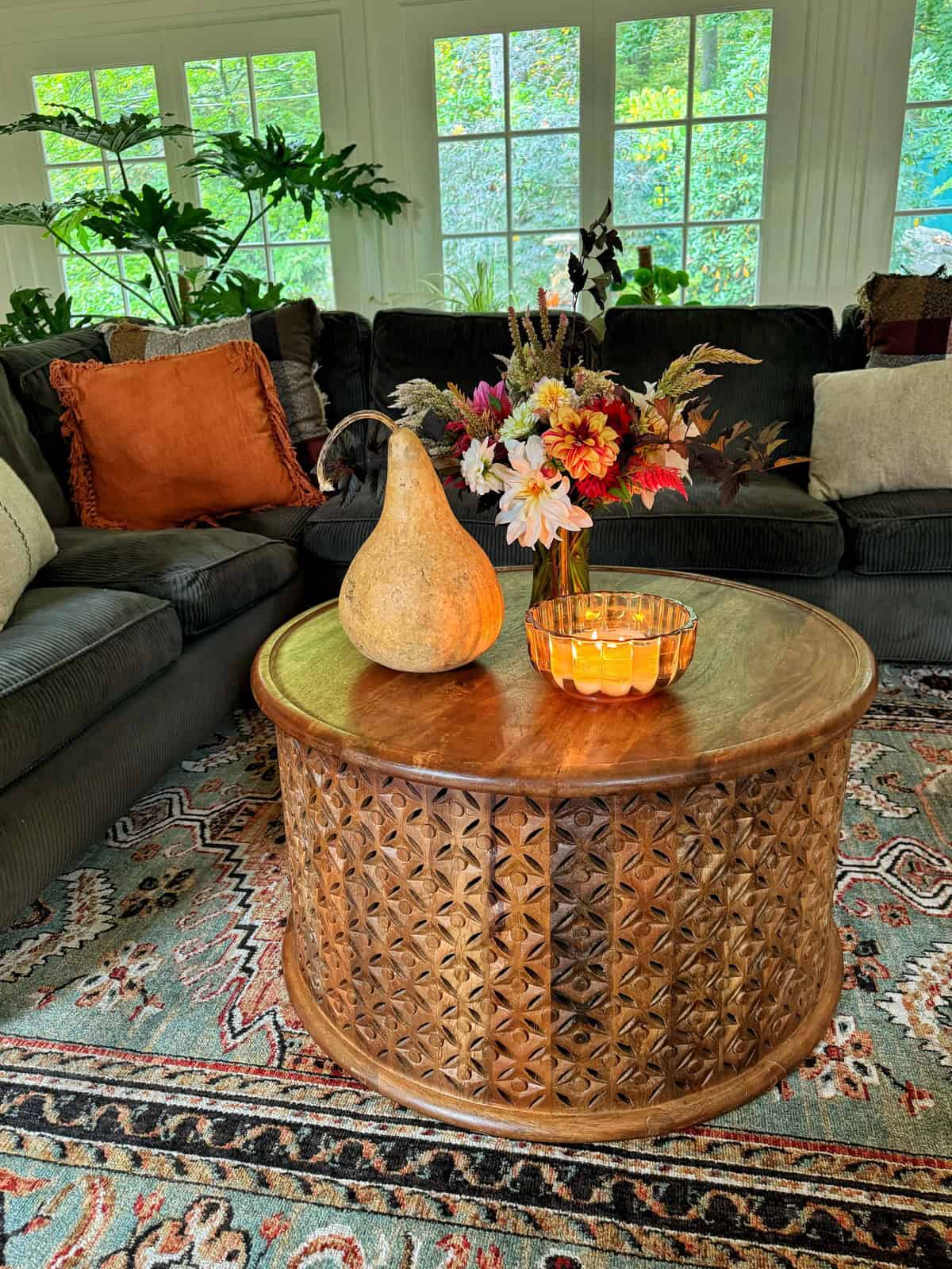 A cozy living room features a carved wooden coffee table with a vase of colorful flowers, a gourd, and a lit candle. The table sits on a patterned rug, surrounded by a dark green sofa with orange and white cushions. Large windows show greenery outside.