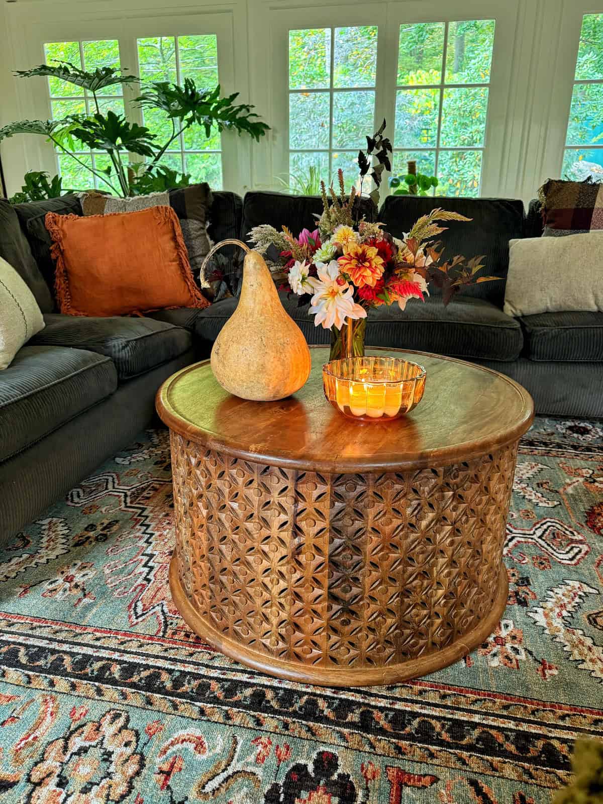 A cozy living room with a dark green sofa, decorated with orange and plaid pillows. A round wooden coffee table features a vase of colorful flowers, a decorative gourd, and an amber glass dish. Large windows in the background show green foliage.