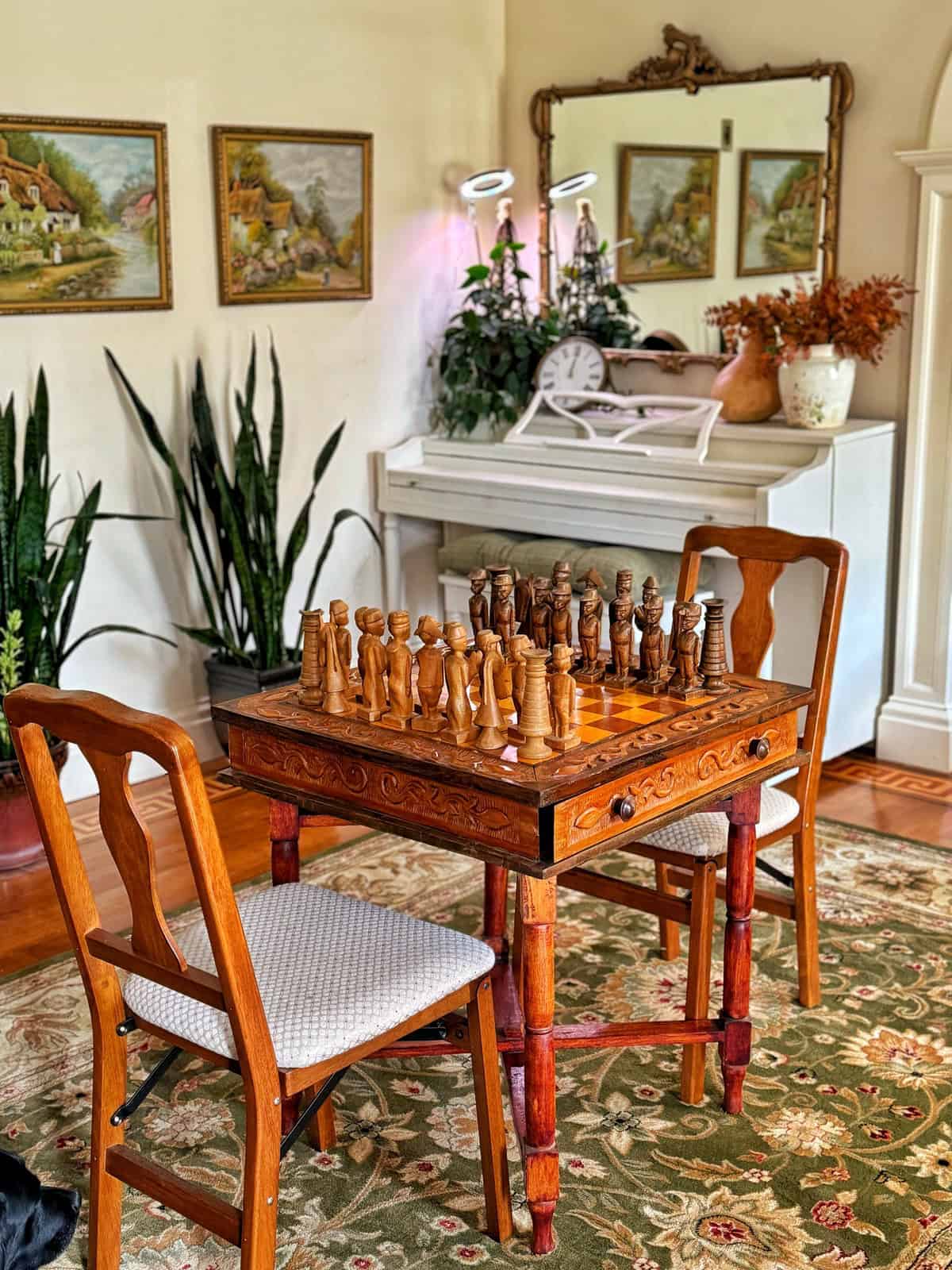 A cozy room with a wooden chessboard set up on a table between two chairs. A white piano, potted plants, and framed landscape paintings adorn the background. A large mirror reflects part of the scene, adding warmth to the space.