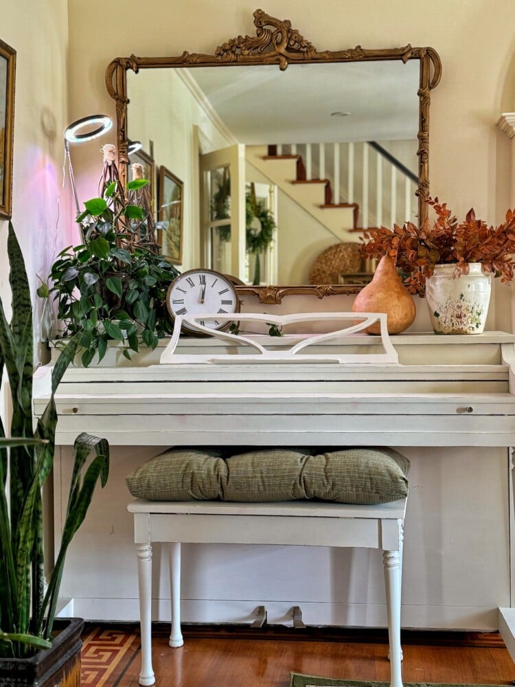 A cozy room with a white piano topped with a decorative clock, potted plants, and autumn-themed decor. An ornate mirror hangs above, reflecting a staircase. A cushioned bench sits in front, and the floor features a patterned rug.