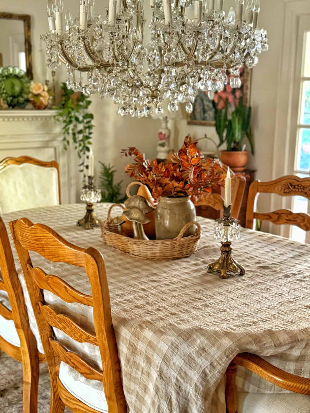 A dining room table with a checkered cloth features a large crystal chandelier above. The table is adorned with a basket containing fall leaves, a ceramic pitcher, and two candlesticks. Wooden chairs surround the table, and a fireplace is visible in the background.