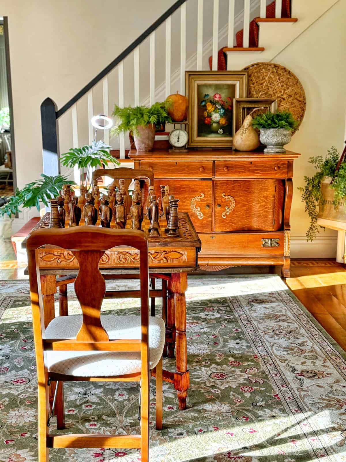 A warmly lit room featuring a wooden chess set on a table with a carved chair. Behind, an ornate cabinet displays plants, framed art, a clock, and decorative items. A patterned rug and staircase are visible in the background.