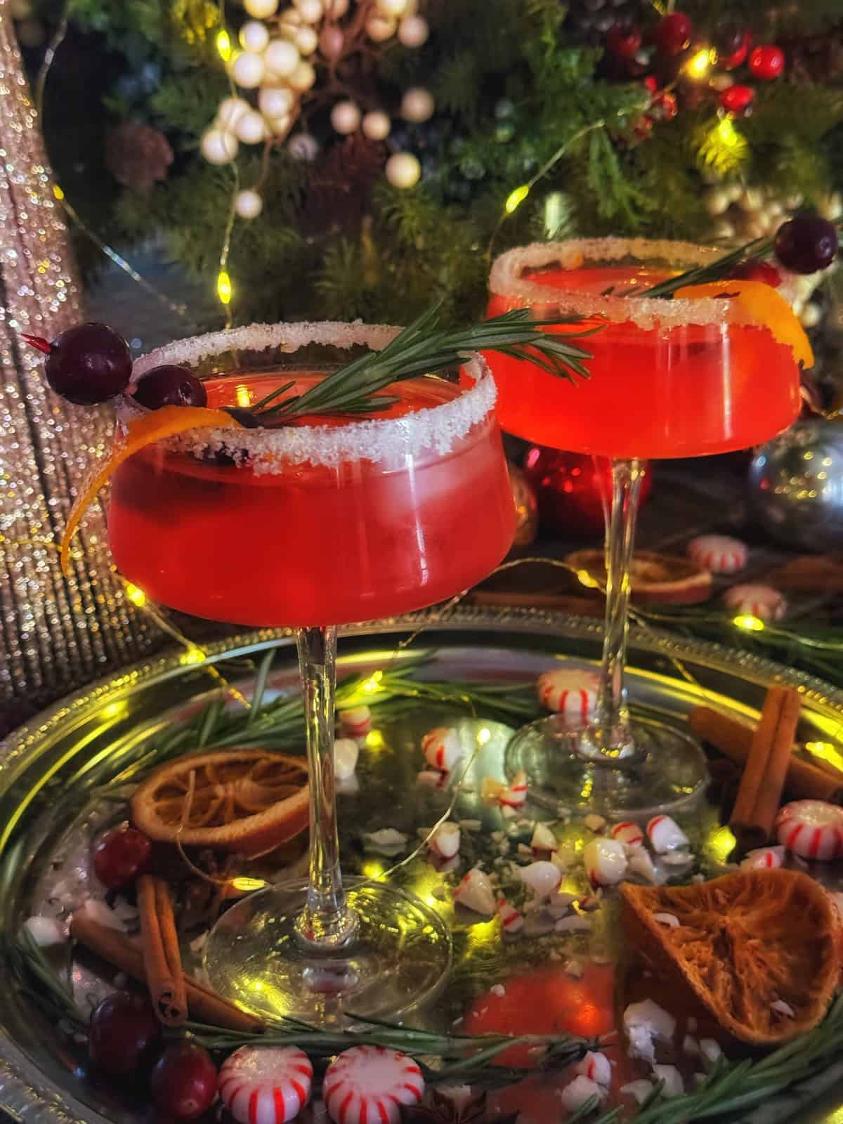 Two festive cocktails with sugared rims and garnished with rosemary and cranberries are on a silver tray. Surrounded by cinnamon sticks, dried orange slices, and peppermint candies, with a background of twinkling lights and greenery.