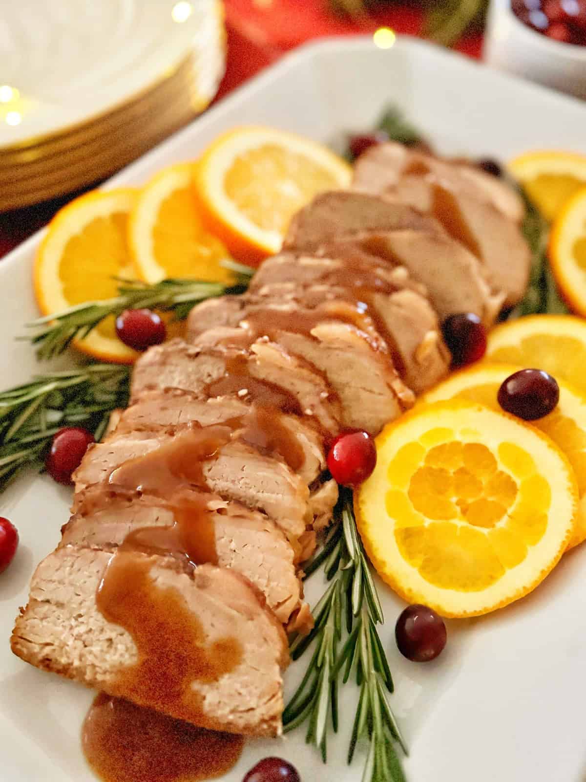Sliced roast pork with gravy is garnished with rosemary sprigs, orange slices, and cranberries, served on a white platter. Plates are visible in the background.