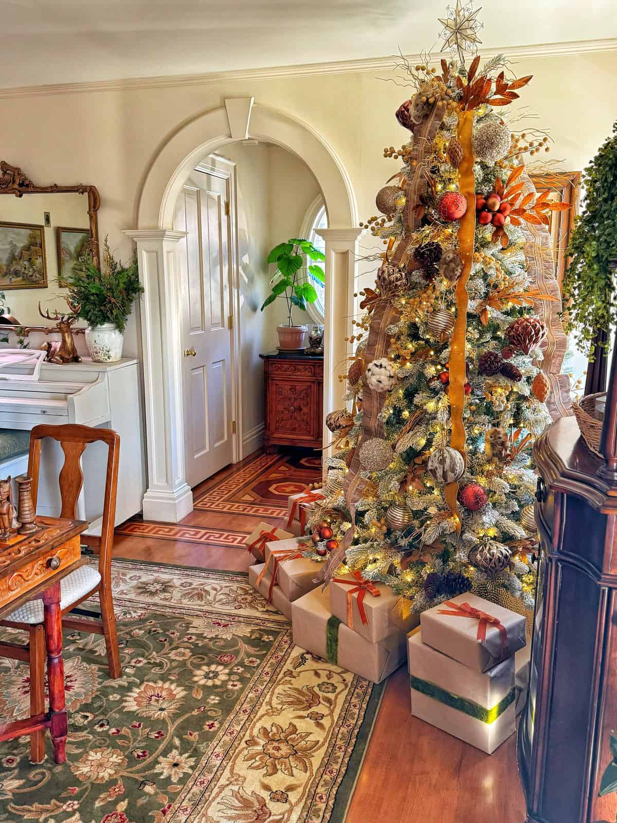 A beautifully decorated Christmas tree stands in a cozy living room, adorned with gold and white ornaments and ribbon. Wrapped presents are placed beneath the tree. Warm lighting enhances the festive atmosphere, with a rug and wooden furniture completing the scene.