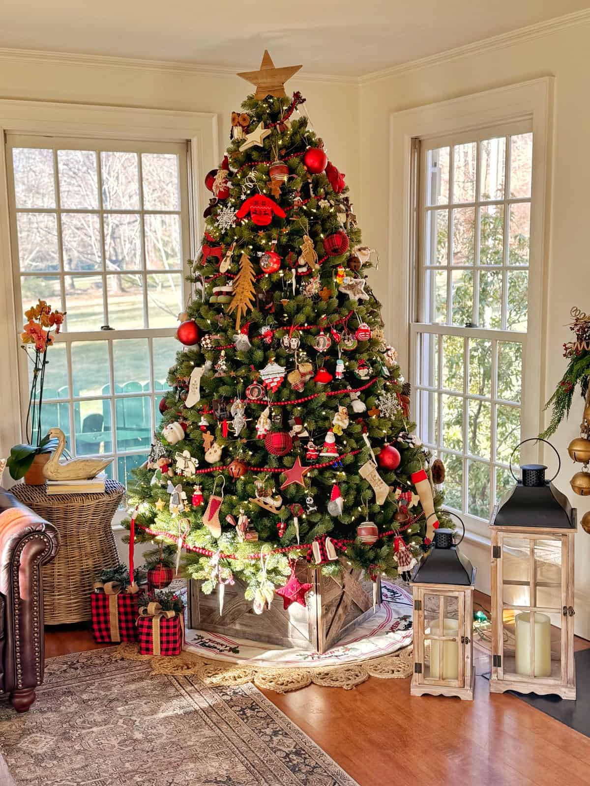 A festive Christmas tree is decorated with red and gold ornaments, ribbons, and a star on top. It is surrounded by lanterns and wrapped gifts. The tree is placed by large windows in a cozy living room with wooden floors.