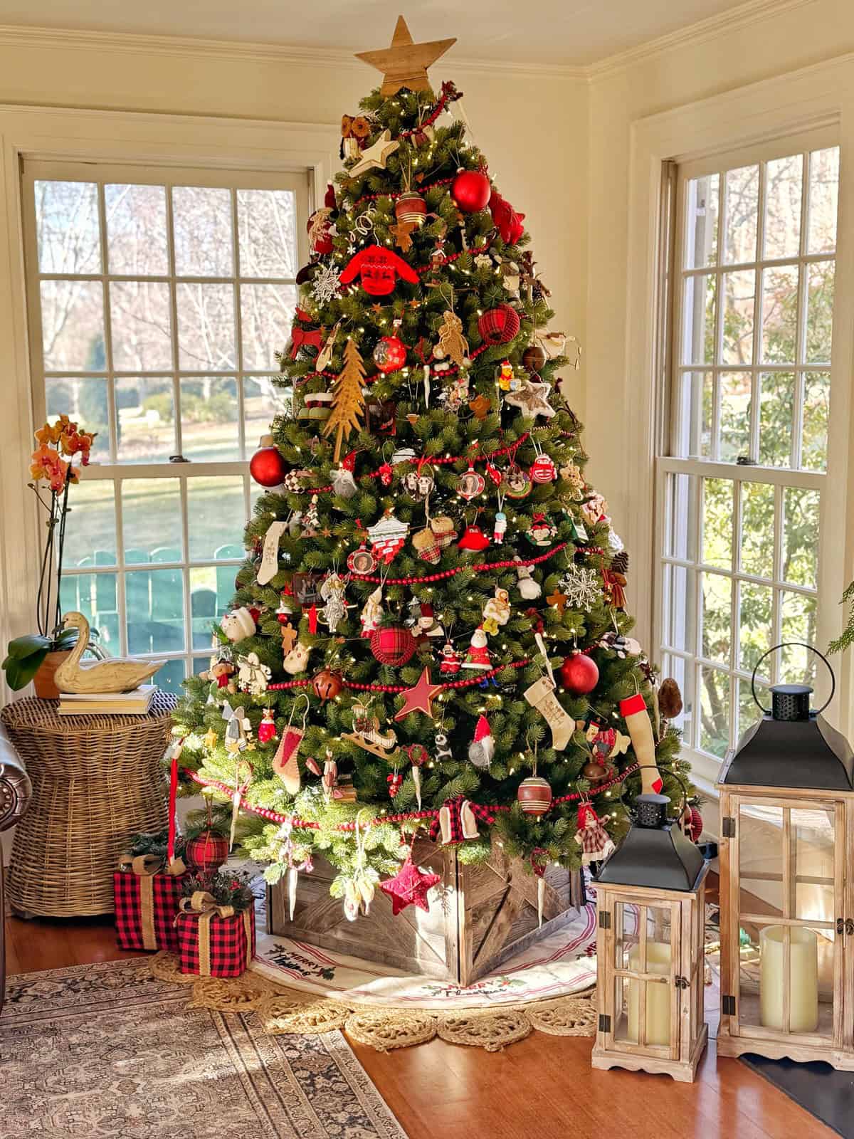A decorated Christmas tree stands in a sunlit room. It is adorned with red and gold ornaments, ribbons, and a star on top. Wrapped presents are placed beneath. Large windows in the background show a view of the outdoors. Lanterns sit on the floor nearby.