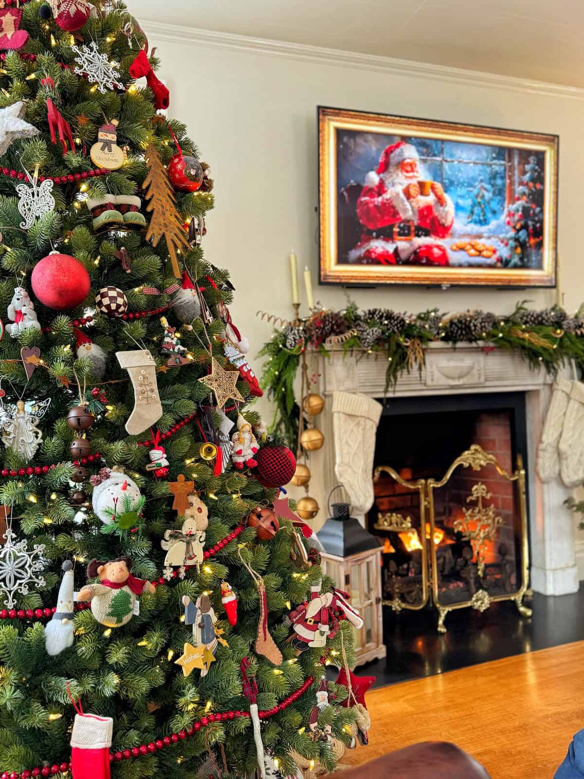 A festive living room with a decorated Christmas tree adorned with ornaments and lights. A fireplace is lit, with stockings hanging above and a garland draped across the mantel. A Santa Claus painting is displayed over the fireplace.