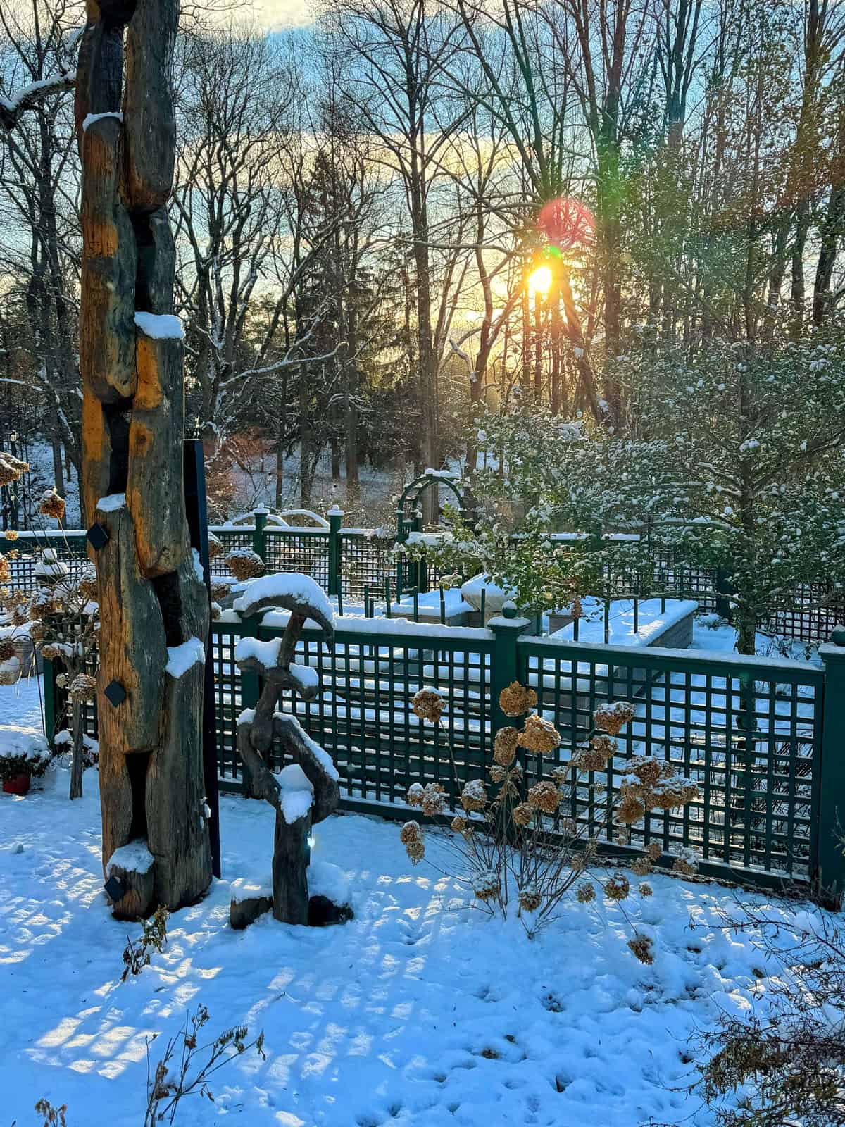 A serene winter garden scene with fresh snow covering the ground and trees. Sunlight filters through the trees, casting warm hues on the white landscape. A wooden sculpture stands amidst the snow, and a trellis fence borders the area.