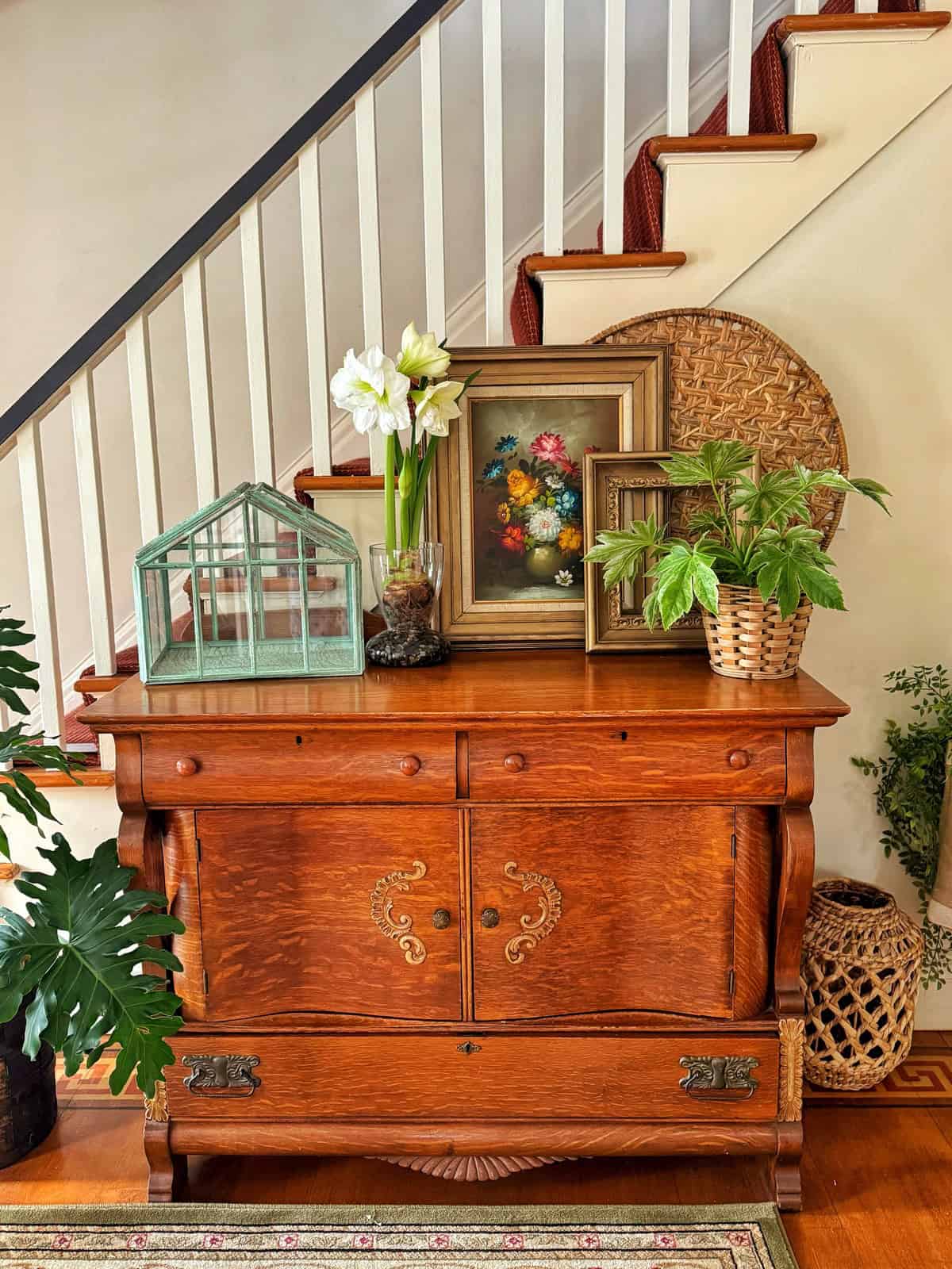 A wooden cabinet stands at the base of a staircase, adorned with framed floral artwork, a small glass terrarium, white flowers in a vase, and potted plants. A woven circular decoration and a plant pot are placed nearby.