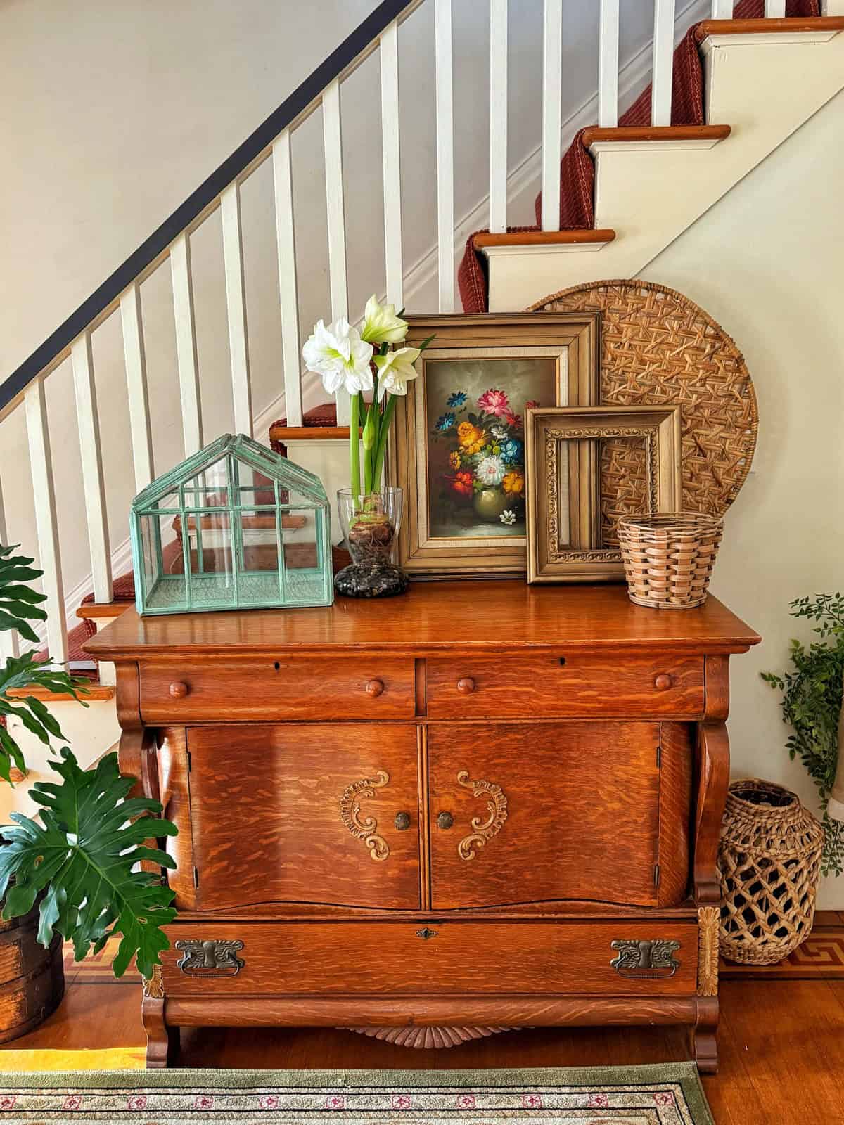 A vintage wooden cabinet sits by a staircase with white railings. On the cabinet are decorative items: a glass terrarium, a potted plant, framed floral art, and baskets. A patterned rug is on the wooden floor, and greenery adds a fresh touch.