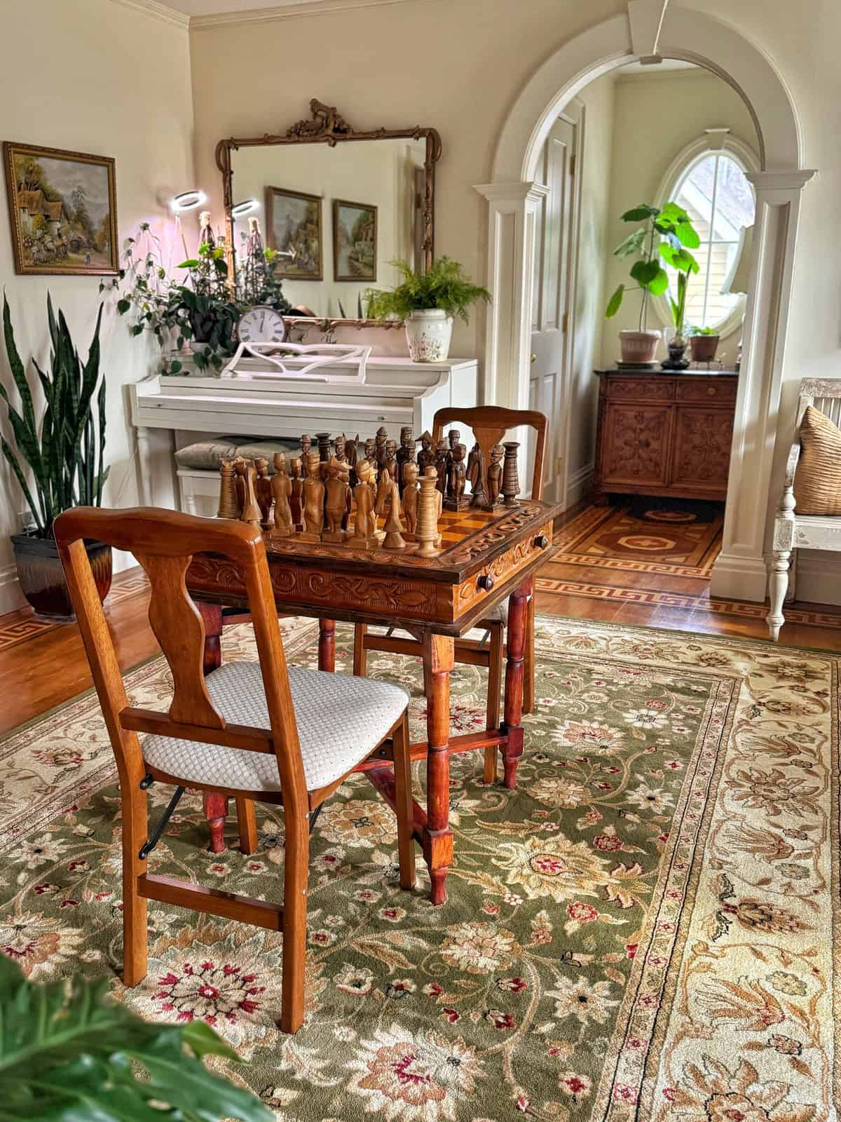 A cozy room with a wooden chess set on a table, surrounded by chairs. The floor is covered with an ornate floral carpet. A white piano and plants decorate the background, with a mirror and paintings on the wall. An arched doorway leads to another area.