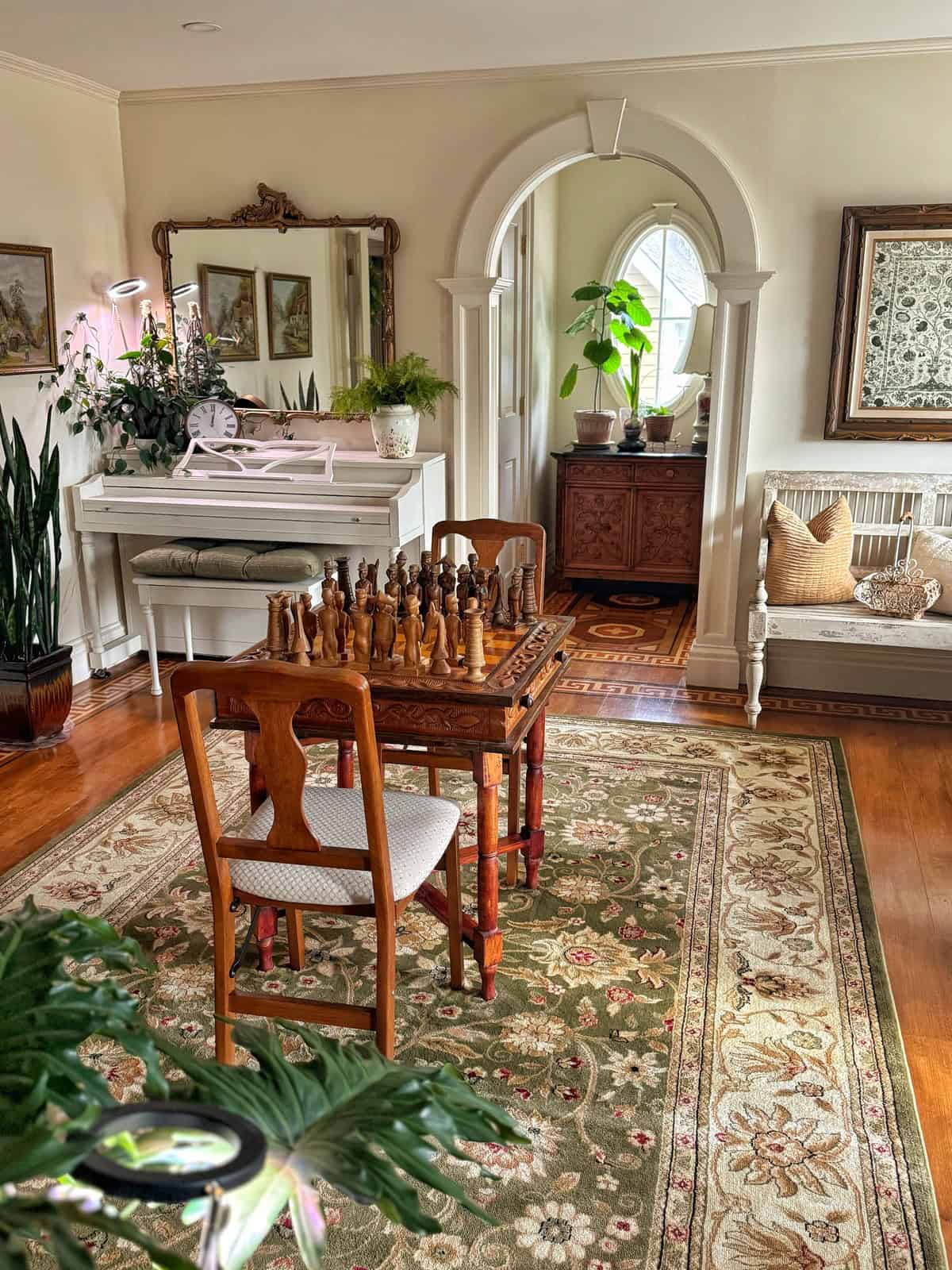 A cozy living room with a white piano, potted plants, and a chess set on a wooden table with chairs. The room has ornate decor, including a large rug, framed art, and a bench with cushions. An arched doorway leads to another area.