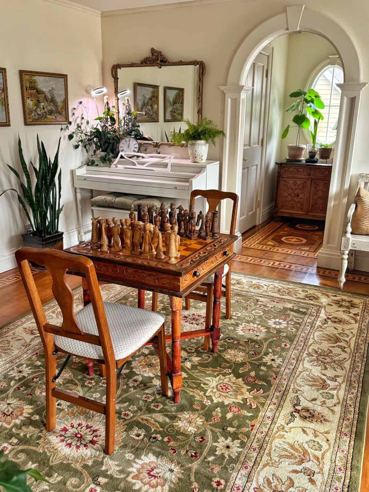 A cozy room with a decorative chess table and chairs on a floral rug. A white piano with plants and framed art is against the wall. An elegant arched doorway leads to another space. Natural light highlights green plants throughout.