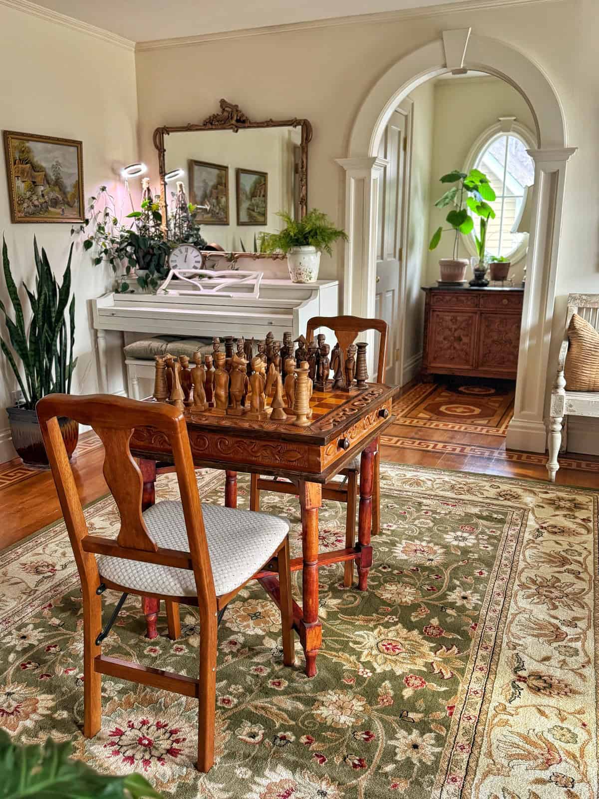 A cozy room features a wooden chess table with matching chairs on a floral rug. A white piano with plants and a mirror sits in the background. Arched doorway leads to another area, with more tropical indoor plants and art on the walls. Natural light fills the space.