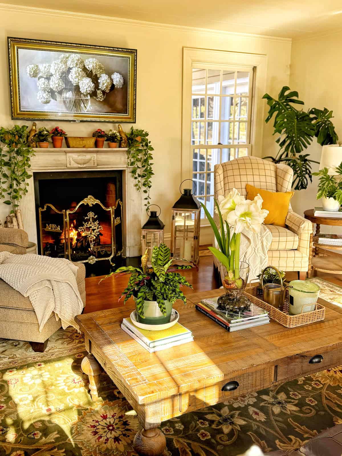 A cozy living room with a lit fireplace, a wooden coffee table, and two armchairs. A framed painting hangs above the mantel, surrounded by tropical indoor plants. Sunlight filters through a window, illuminating the room and casting shadows on the floor.