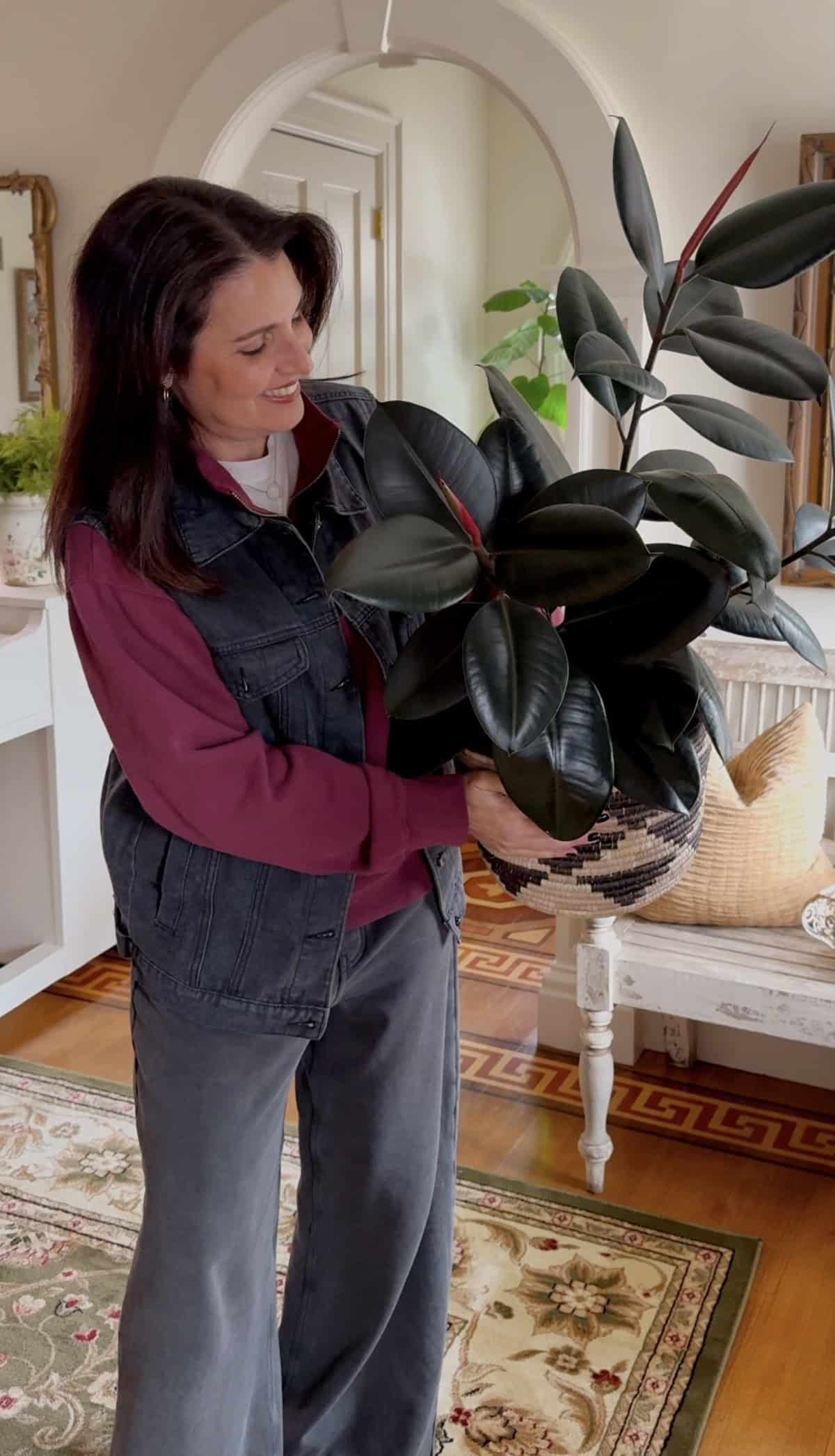 A woman with long dark hair, wearing a maroon shirt and denim vest, is holding a large potted rubber plant in a living room. The room has wooden flooring, a woven basket, and light filtering through a window.