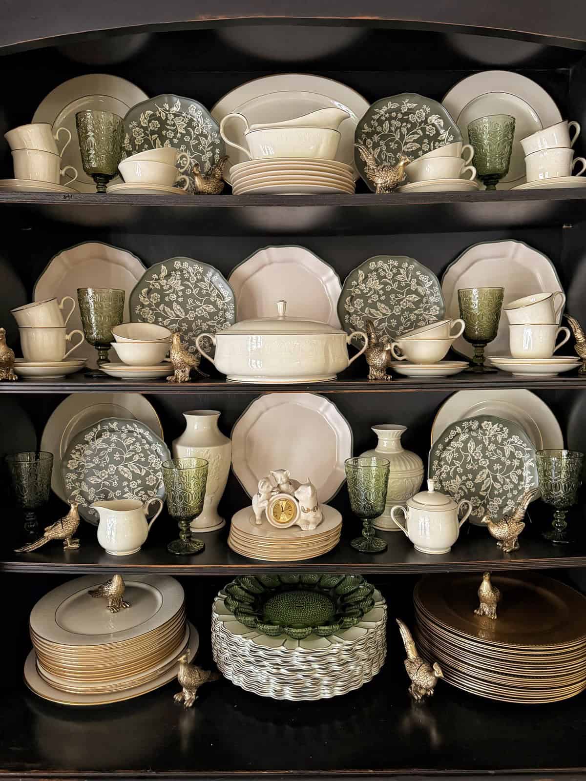 A neatly arranged cabinet displays ceramic plates, bowls, and cups in white and green floral patterns. Green goblets and decorative rooster figurines add charm. Gold-rimmed plates and intricate detailing complete the elegant setup.