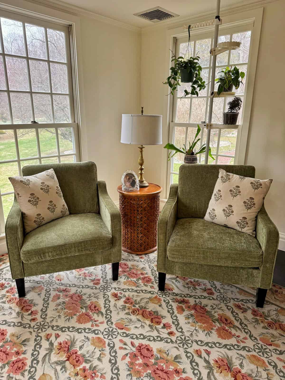 Two green armchairs with leaf-patterned pillows facing a wooden side table with a lamp and small framed photo. Behind them are large windows and hanging plants, all set on a floral patterned rug.