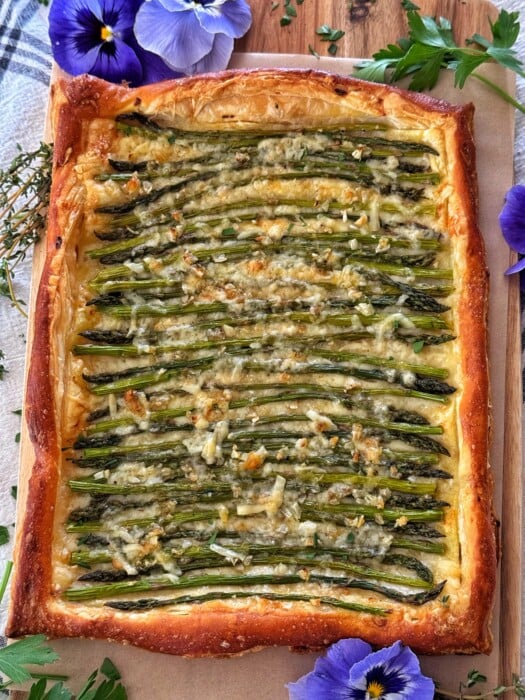 A rectangular asparagus tart with neatly arranged spears is topped with a sprinkling of cheese. This delightful creation is garnished with purple flowers and fresh herbs, all elegantly displayed on a rustic wooden surface.
