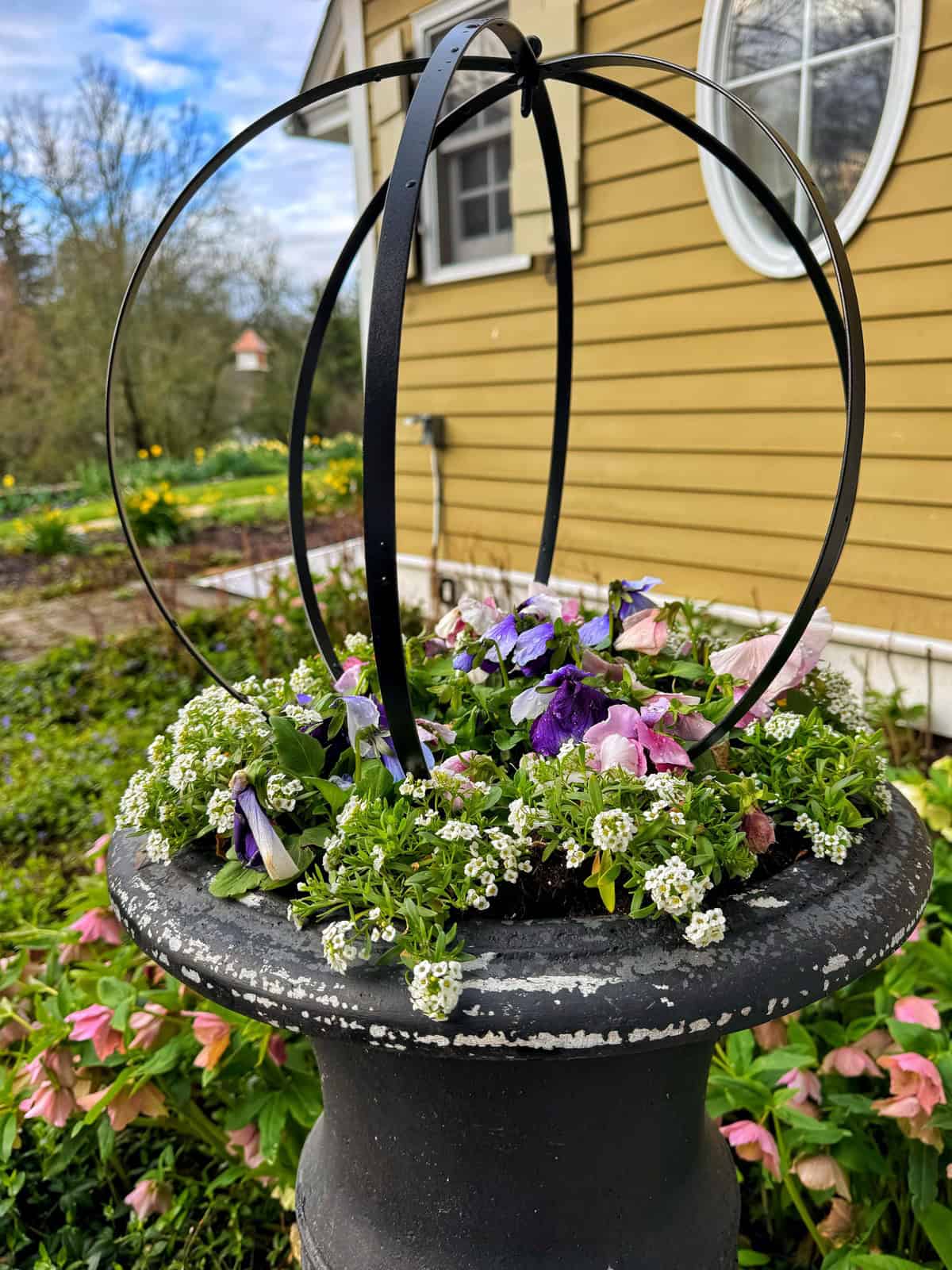 A black metal planter with blooming purple, pink, and white flowers sits in front of a yellow house with white trim and a round window. The garden in the background is lush and green.