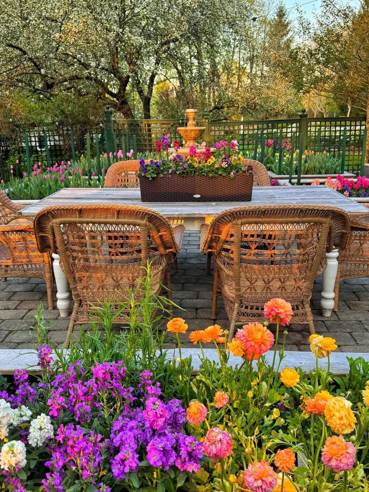 Wicker chairs surround a white table on a stone patio, with colorful flowers in the foreground and a planter centerpiece. Lush green trees and blooming flowers fill the background, evoking a serene garden setting.