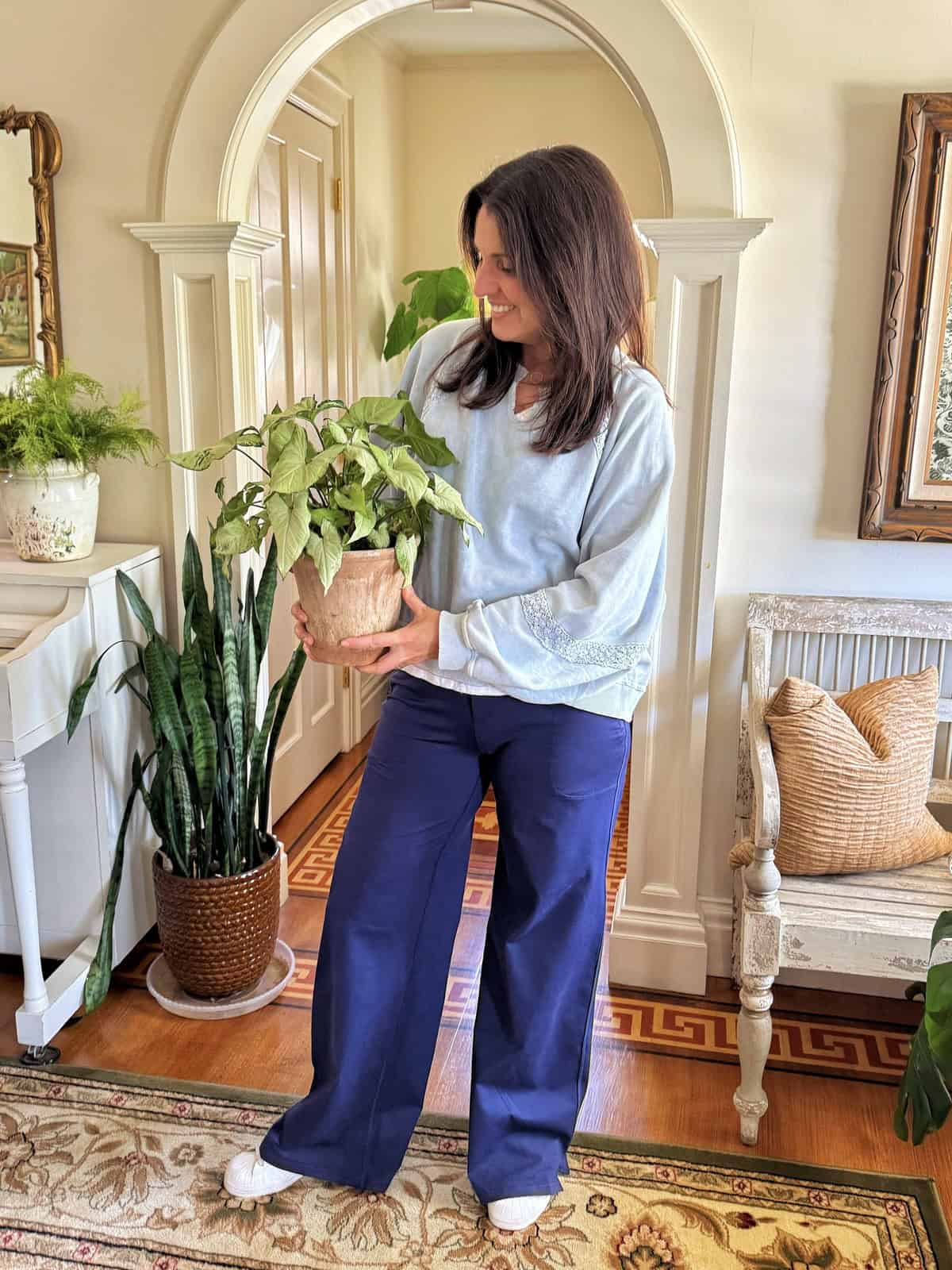 A woman with long dark hair, wearing a light blue top and blue pants, stands indoors holding a potted plant. She is smiling and surrounded by houseplants, with sunlight filling the room decorated in neutral tones.