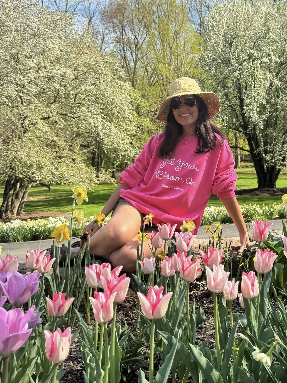 A woman in a pink sweatshirt, sunglasses, and a straw hat sits among blooming tulips and daffodils in a sunny garden, smiling at the camera. Lush green trees and blue sky are in the background.