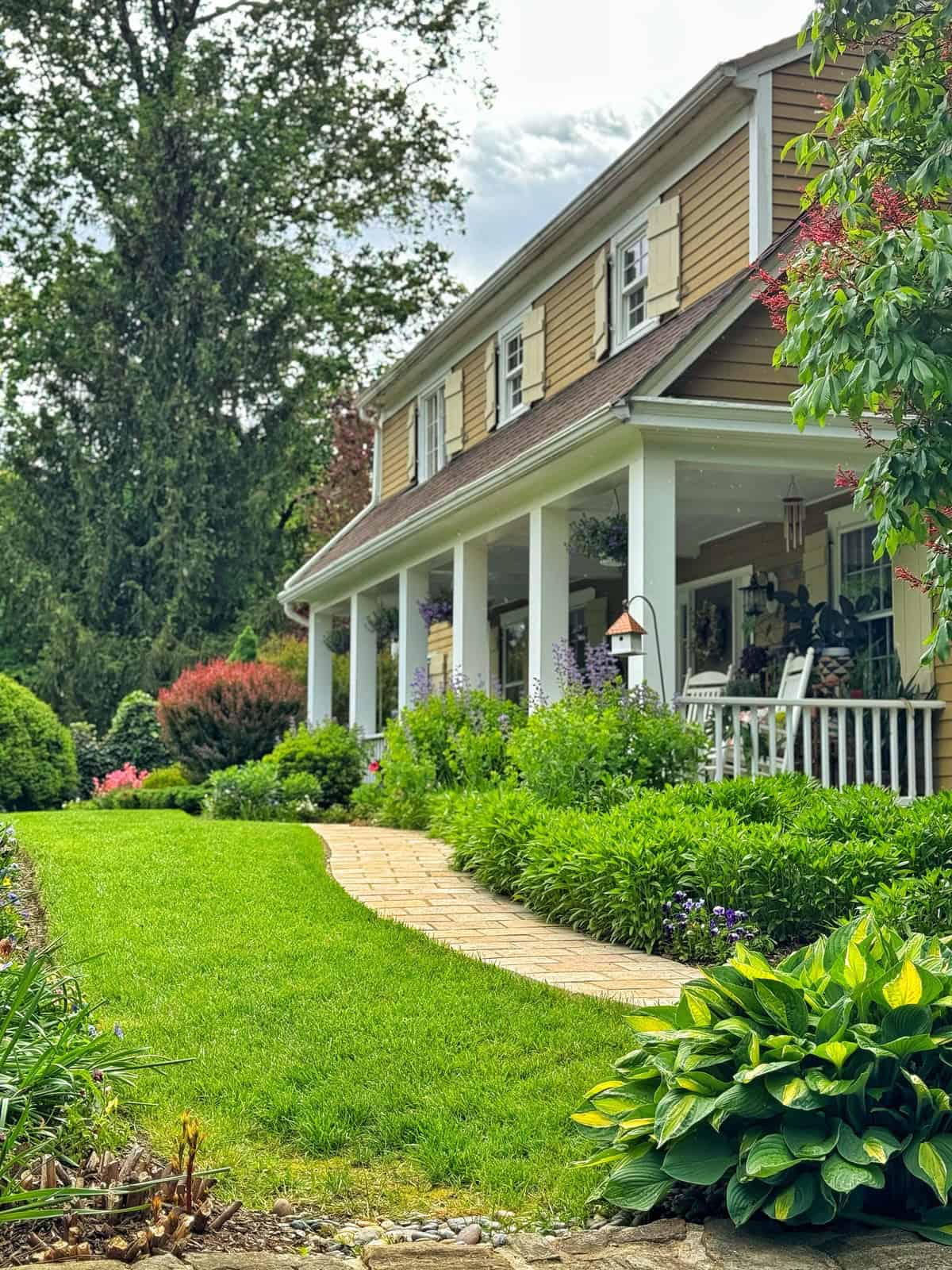 A well-maintained house with tan siding and white trim, a covered front porch with rocking chairs, and a brick path winding through a lush, green garden with various shrubs and flowering plants.