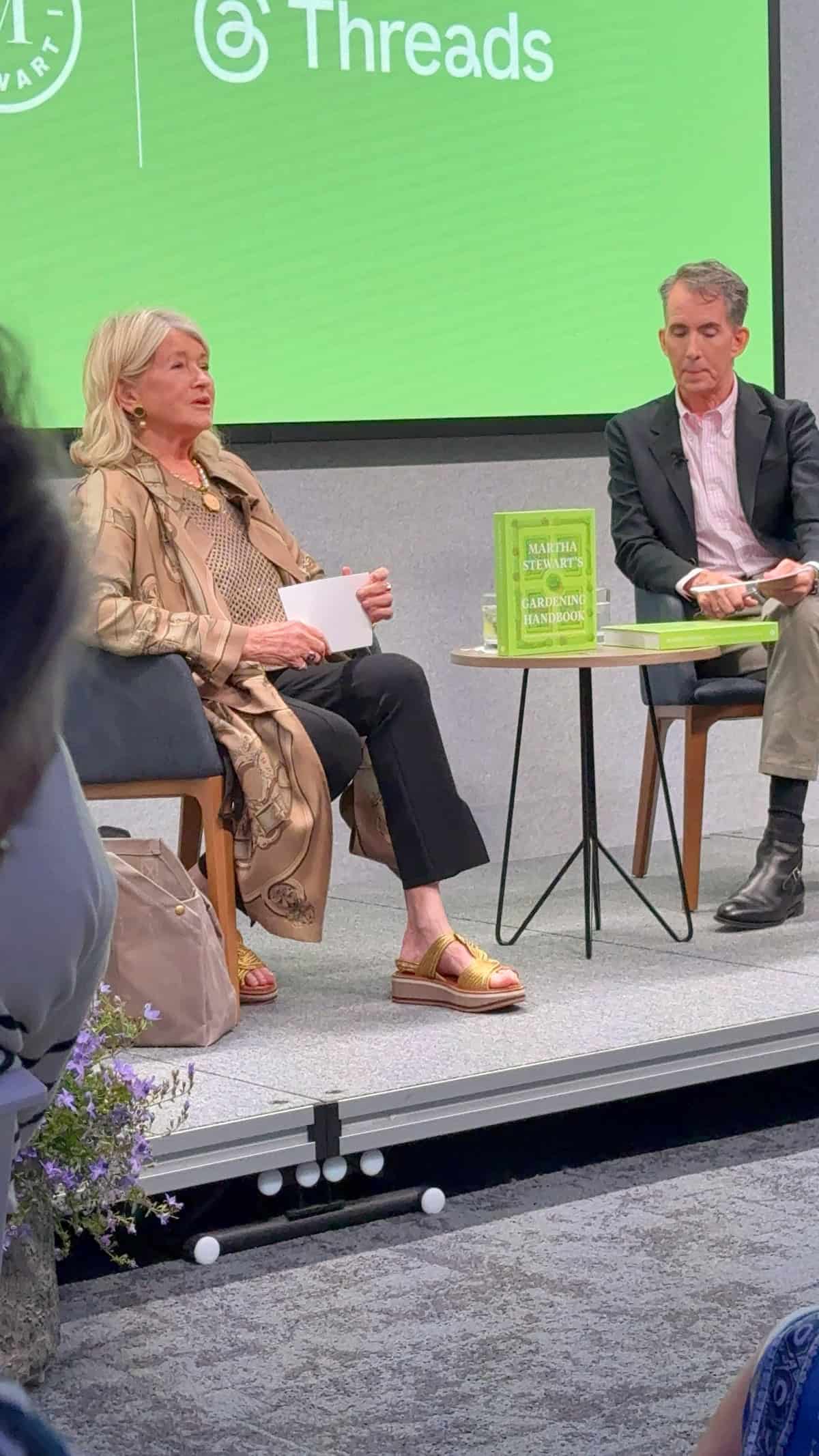 Two people (martha stewart and kevin) sit on stage during a discussion. The woman on the left speaks while holding a card; the man on the right listens. A green book is displayed on the table between them, with a green "Threads" backdrop behind.