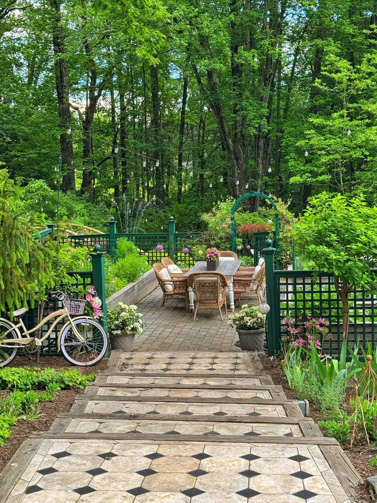 Stone steps lead to a cozy garden patio with wicker chairs and a table, surrounded by lush greenery, flowering plants, a bicycle, and a green trellis. Tall trees create a peaceful, inviting outdoor setting.