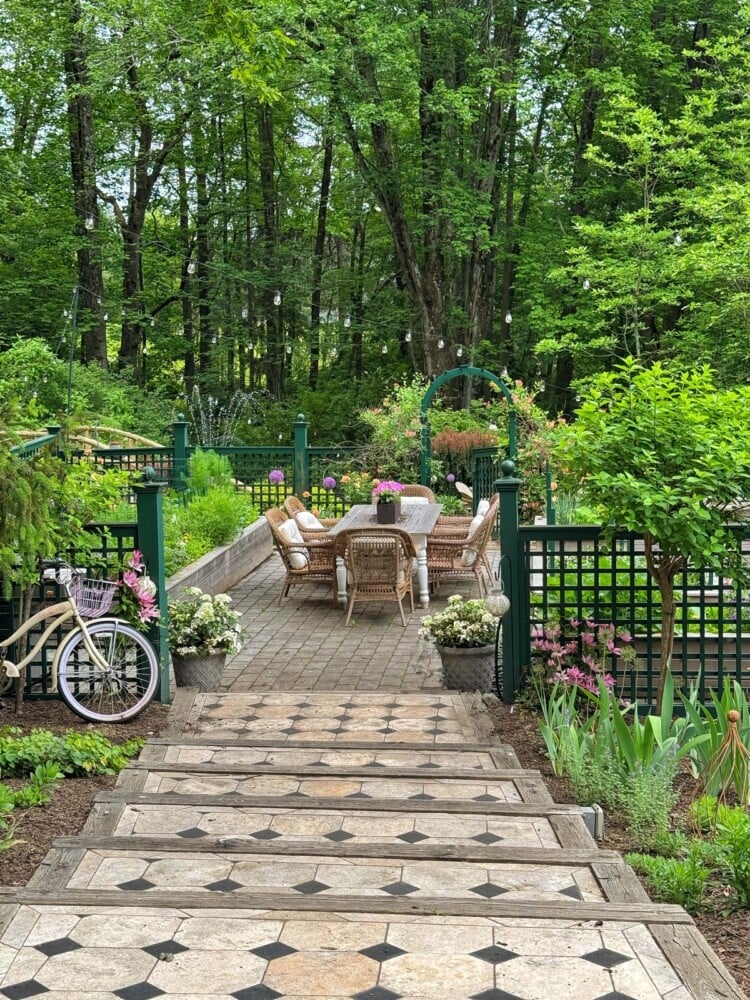 A stone pathway leads down steps to a potager garden patio with wicker chairs and a table, surrounded by green trees, flowers, and string lights. A white bicycle with a basket is parked by a green lattice fence.