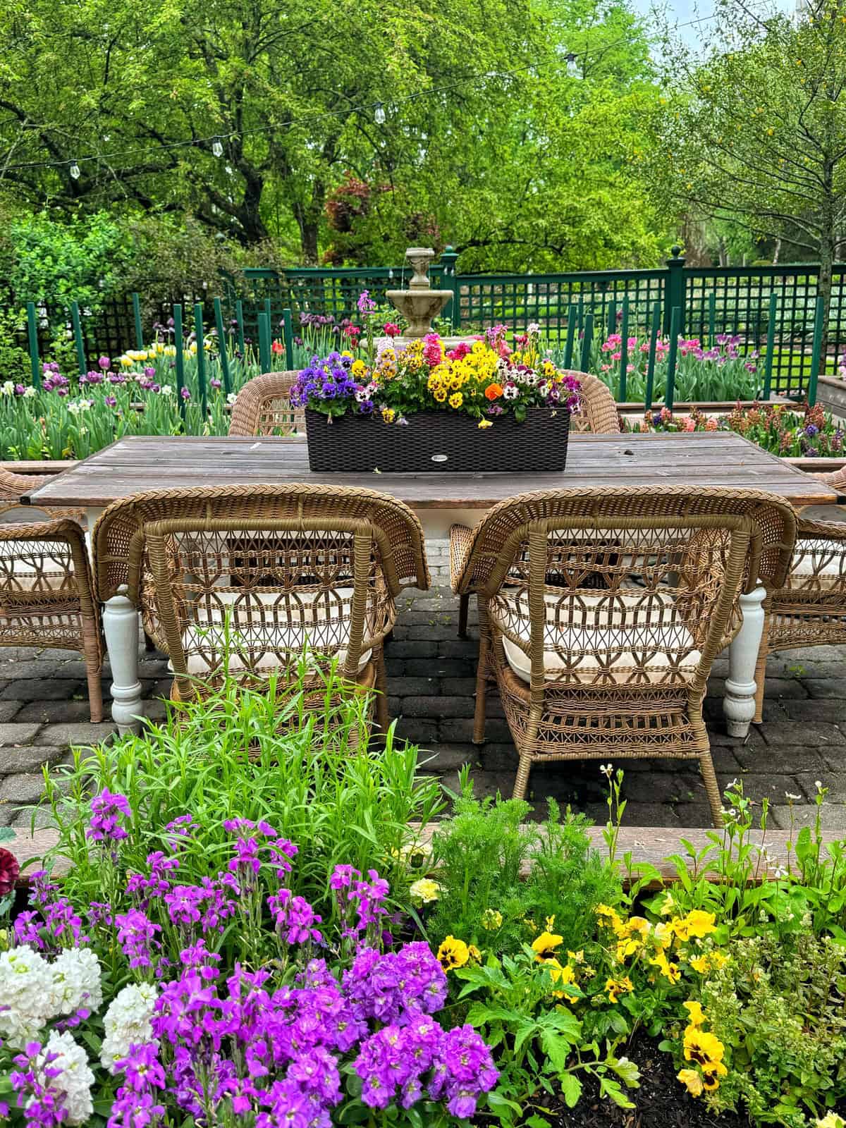 A wooden outdoor table with wicker chairs sits on a stone patio surrounded by blooming flowers, with a large planter of colorful flowers on the table and lush green trees in the background.