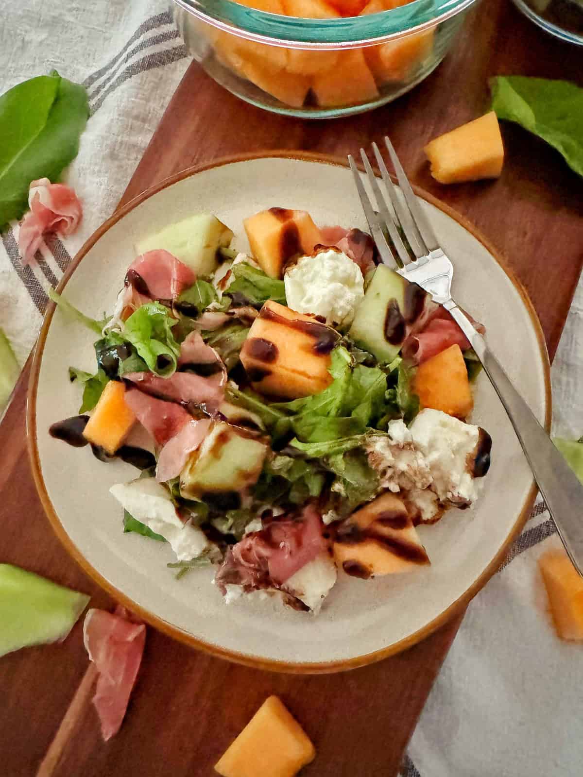 A plate of salad with green lettuce, prosciutto, melon cubes, mozzarella, and balsamic glaze. A fork rests on the plate, and more melon and prosciutto pieces are scattered nearby on a wooden board.