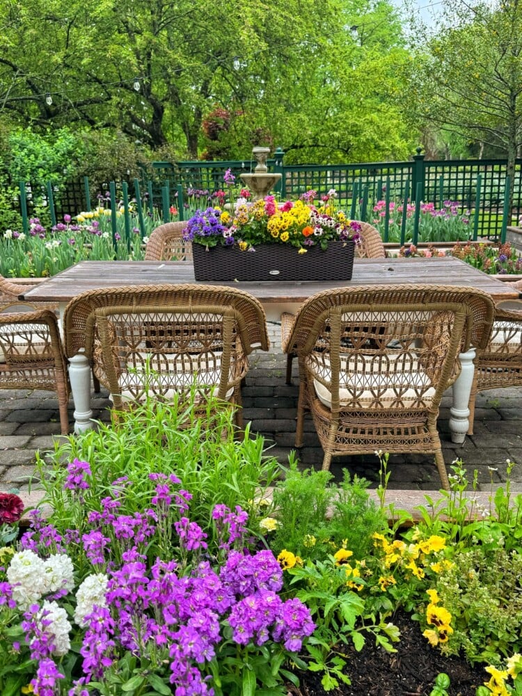 A wicker outdoor dining set with cushions sits on a patio, surrounded by colorful blooming flowers and lush greenery. A basket of vibrant flowers decorates the center of the wooden table.