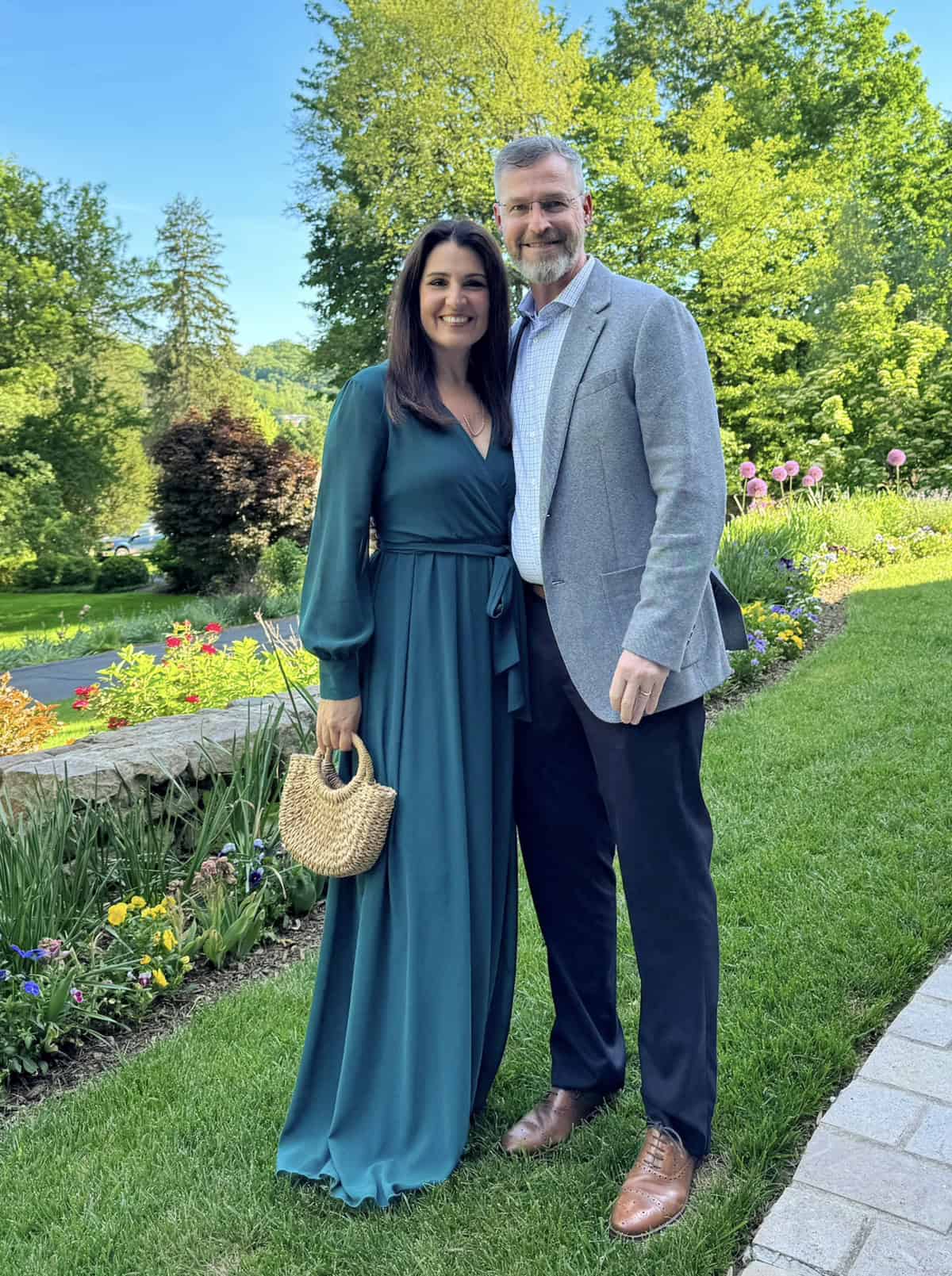 A smiling couple stands on grass in a garden. The woman wears a long teal dress and holds a woven bag; the man wears a gray blazer, white shirt, and brown shoes. Colorful flowers and green trees fill the background.