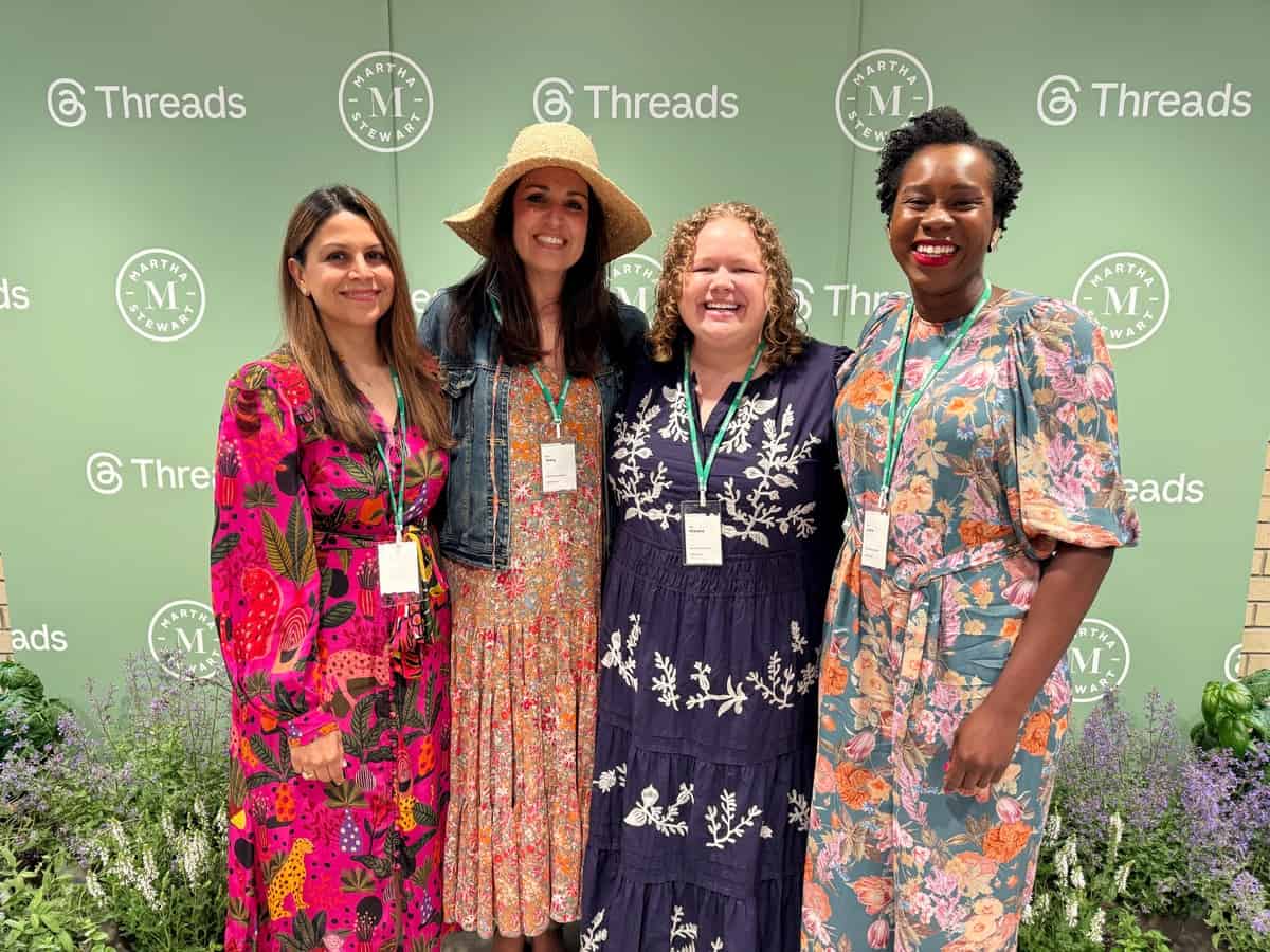 Four women stand smiling together in front of a green backdrop with "Threads" and circular "M" logos, all wearing colorful, patterned dresses and event badges. Flowers and greenery are visible at their feet.