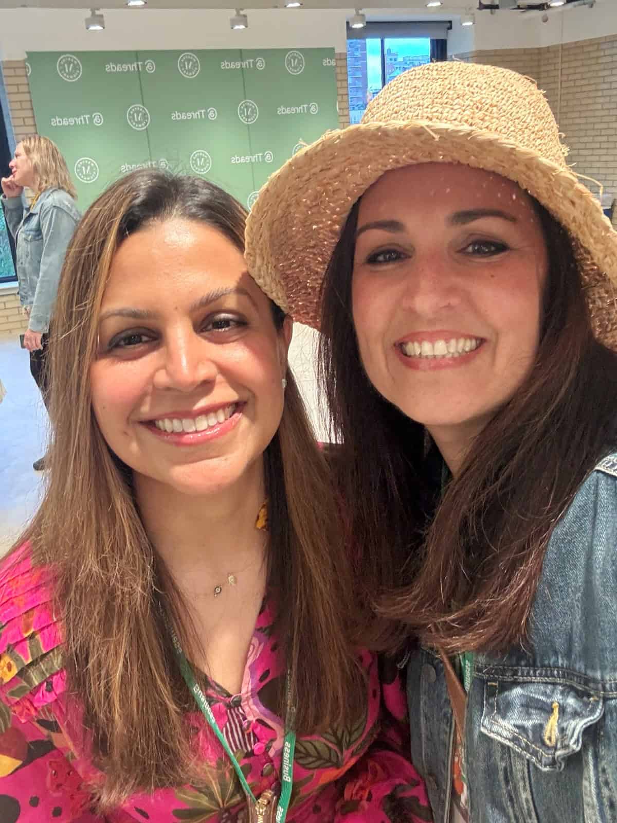 Two women smiling for a selfie indoors. One is wearing a pink patterned top, and the other is wearing a straw hat and denim jacket. A green backdrop with "Threads" logos is visible behind them.