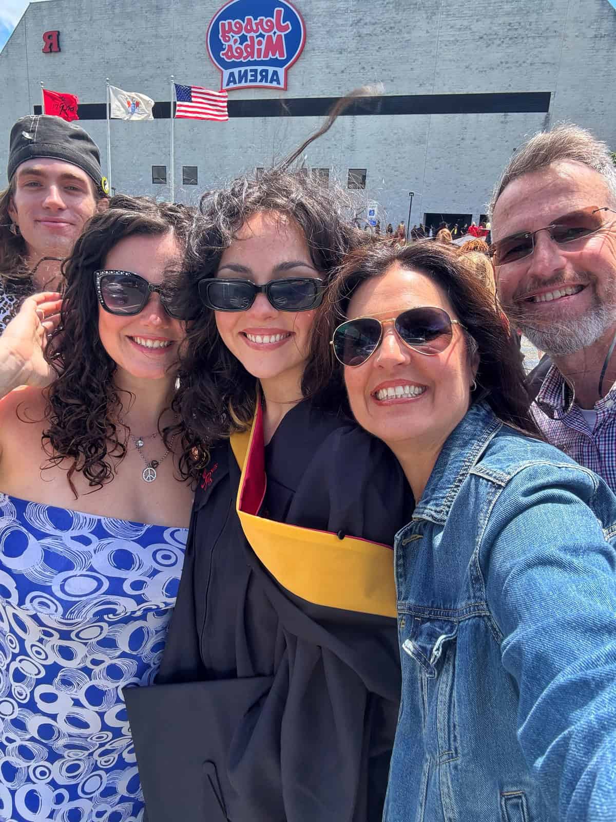 A group of five people smiling and taking a selfie at an outdoor graduation ceremony, with one person wearing a cap and gown. The background shows flags and a building labeled "Jersey Mike's Arena.