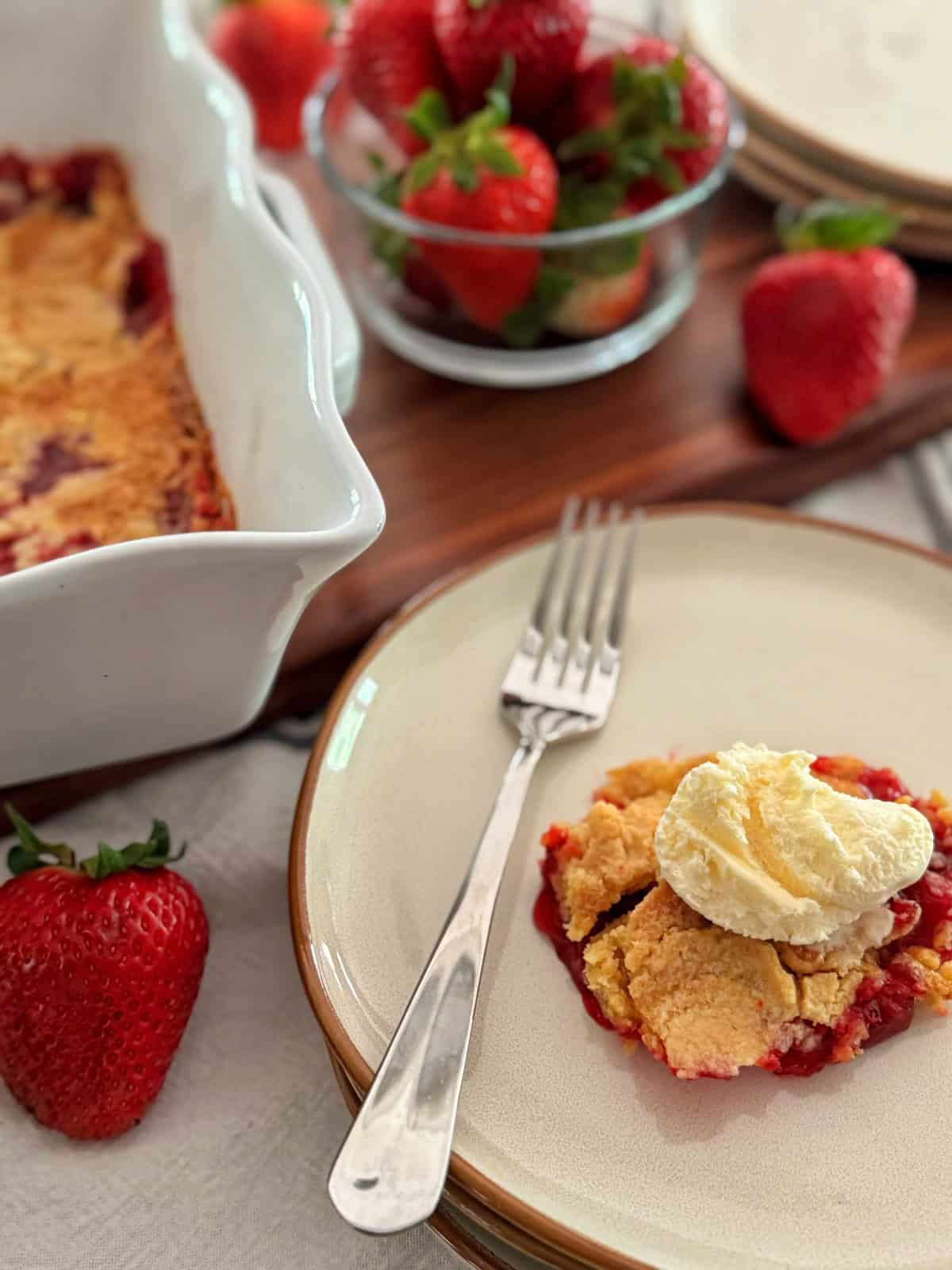 A plate with a serving of strawberry dump cake recipe topped with a scoop of vanilla ice cream, a fork beside it. In the background are fresh strawberries in a bowl and a baking dish with more cobbler.