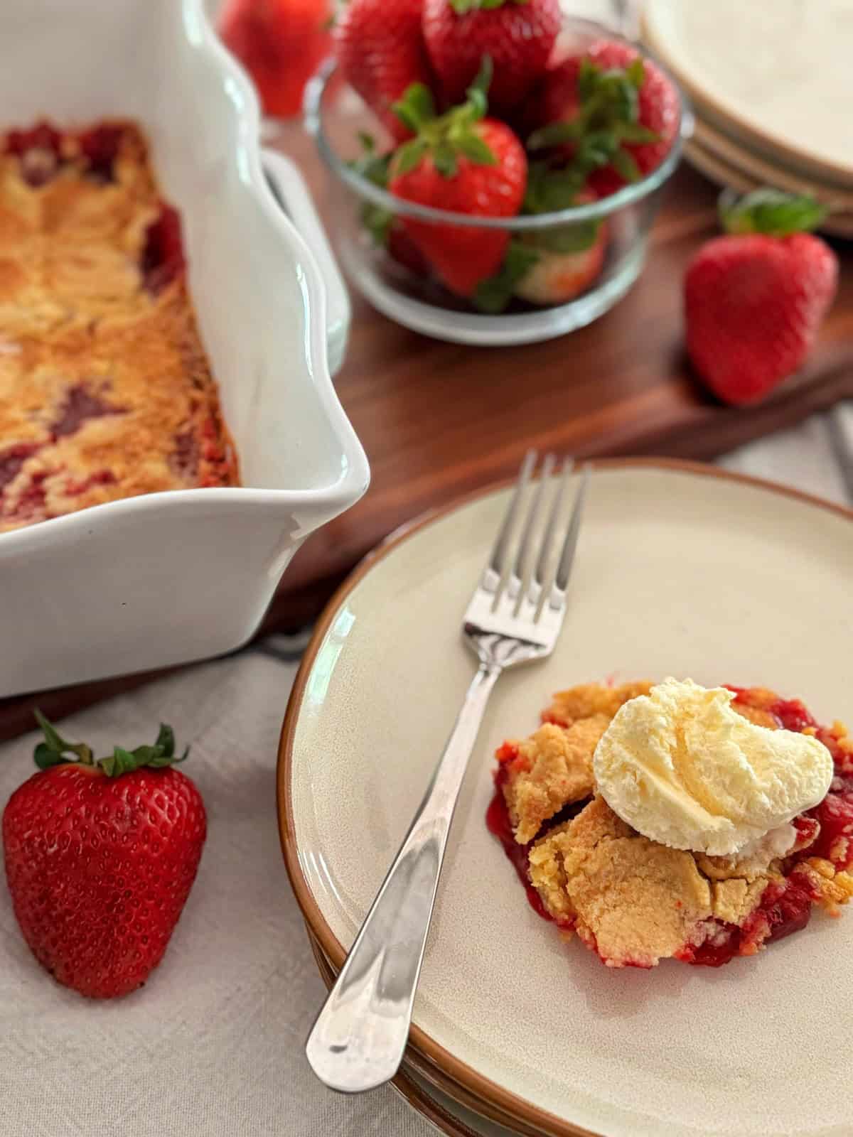 A serving of strawberry dump cake topped with a scoop of vanilla ice cream sits on a plate with a fork. Nearby are fresh strawberries, a bowl of strawberries, and a baking dish of cobbler on a wooden board.