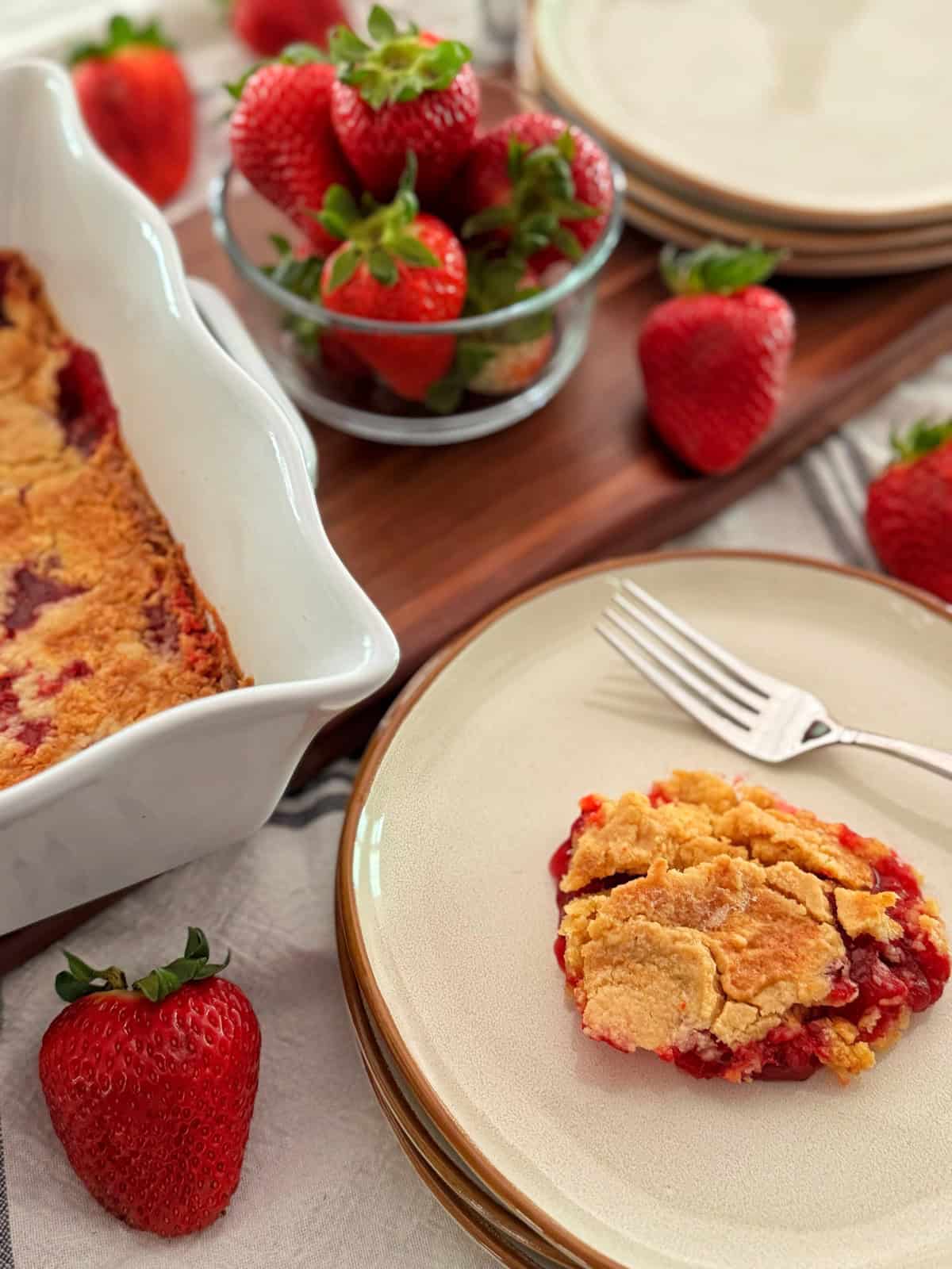 A slice of strawberry dump cake on a plate with a fork, surrounded by fresh strawberries. In the background, a baking dish with more cobbler and a glass bowl of strawberries sit on a wooden tray.