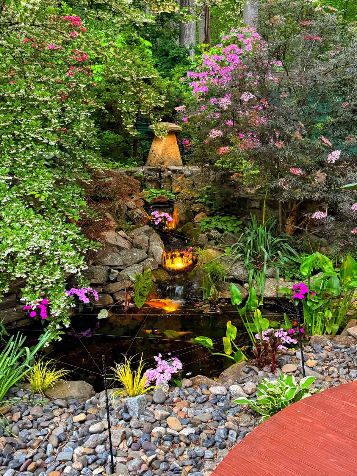 A small garden pond with blooming pink and white flowers, surrounded by rocks and greenery. A waterfall flows into the pond, lit by warm yellow lights, creating a peaceful and vibrant outdoor scene.