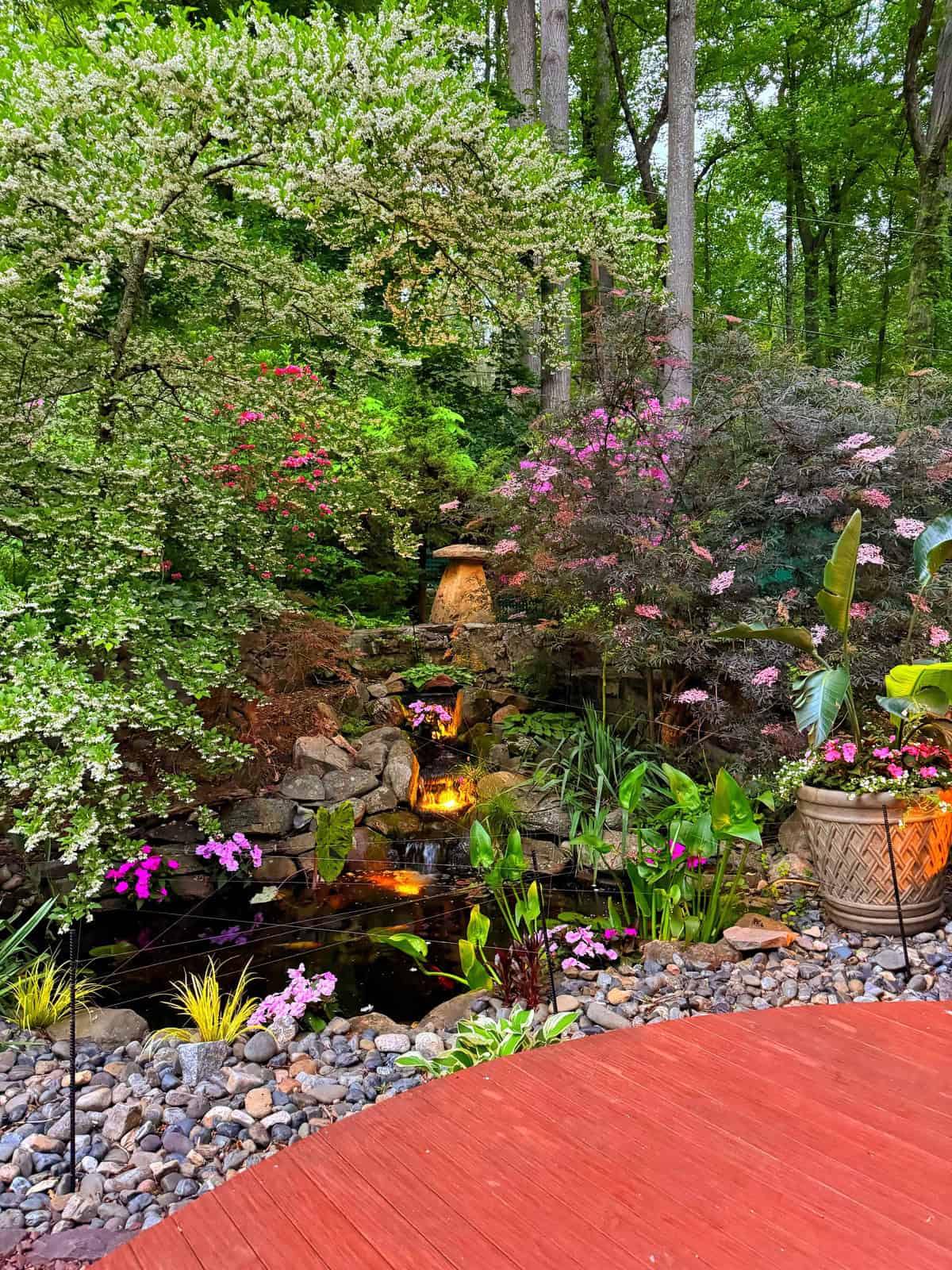 A lush garden with blooming flowers, green trees, a small waterfall flowing over rocks into a pond, and potted plants on a wooden deck. Soft lights illuminate the waterfall and surrounding foliage.
