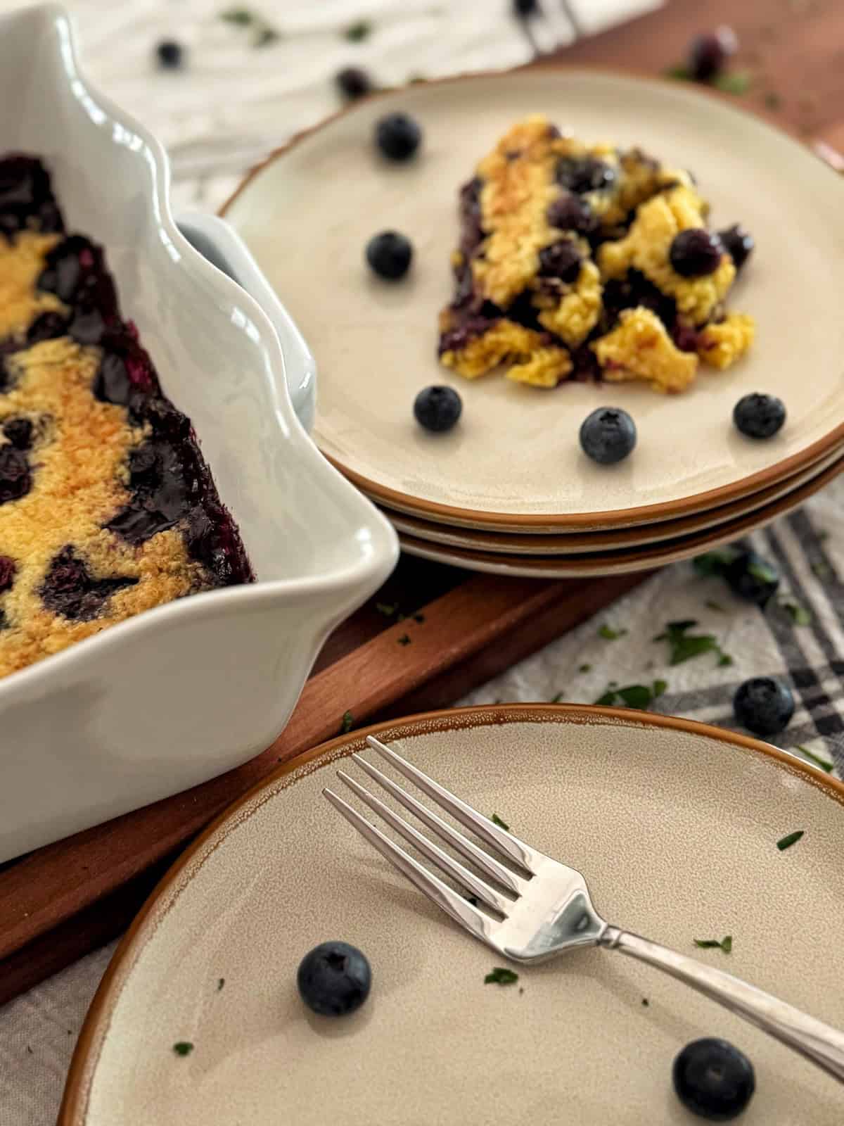 A serving of blueberry dump cake sits on a stack of beige plates next to a ceramic baking dish with more cobbler. Fresh blueberries are scattered around, and a fork rests on the nearest plate.