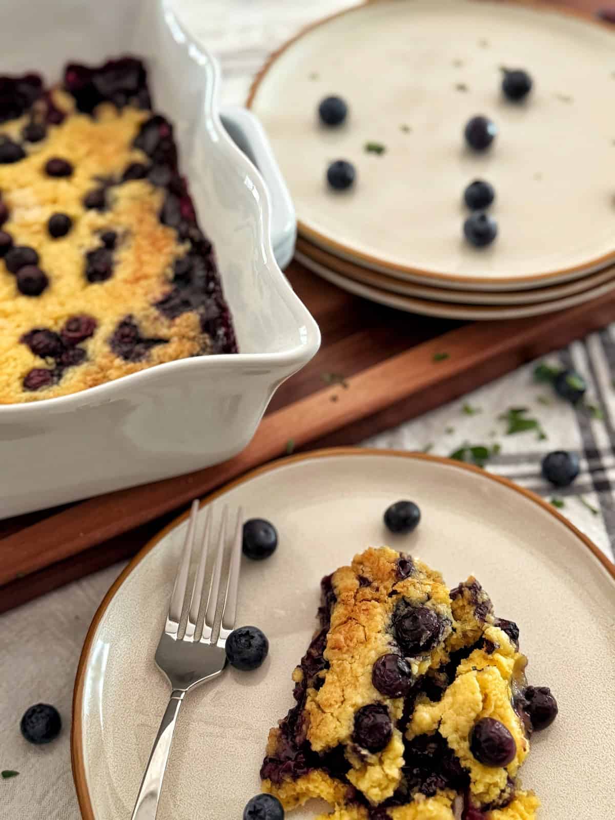 A slice of blueberry dump cake recipe on a plate with a fork, next to a baking dish filled with cobbler and several stacked plates. Fresh blueberries are scattered on the plates and table.