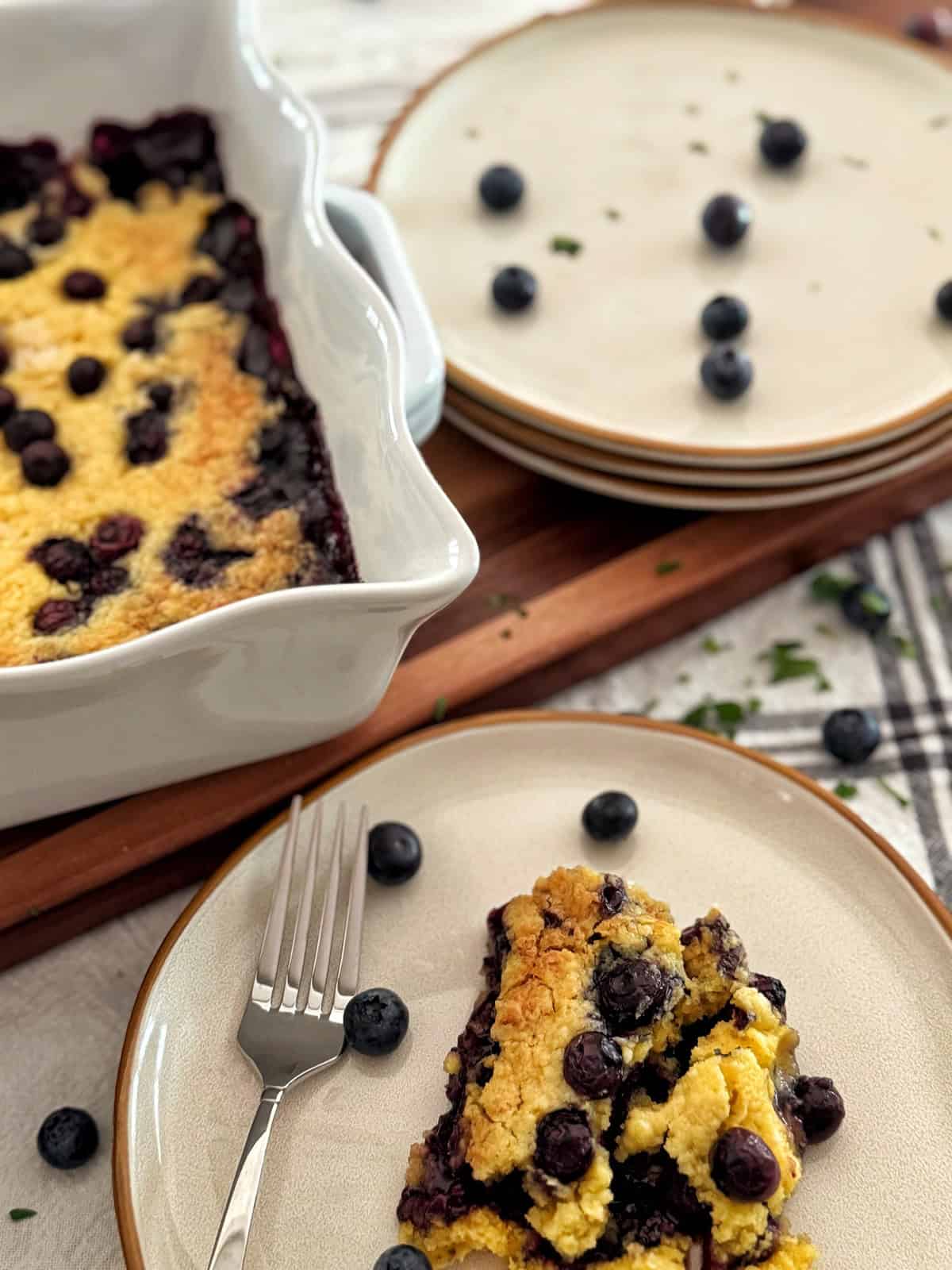 A slice of blueberry dump cake on a plate with a fork and scattered blueberries, next to a baking dish of cobbler and stacked plates in the background.