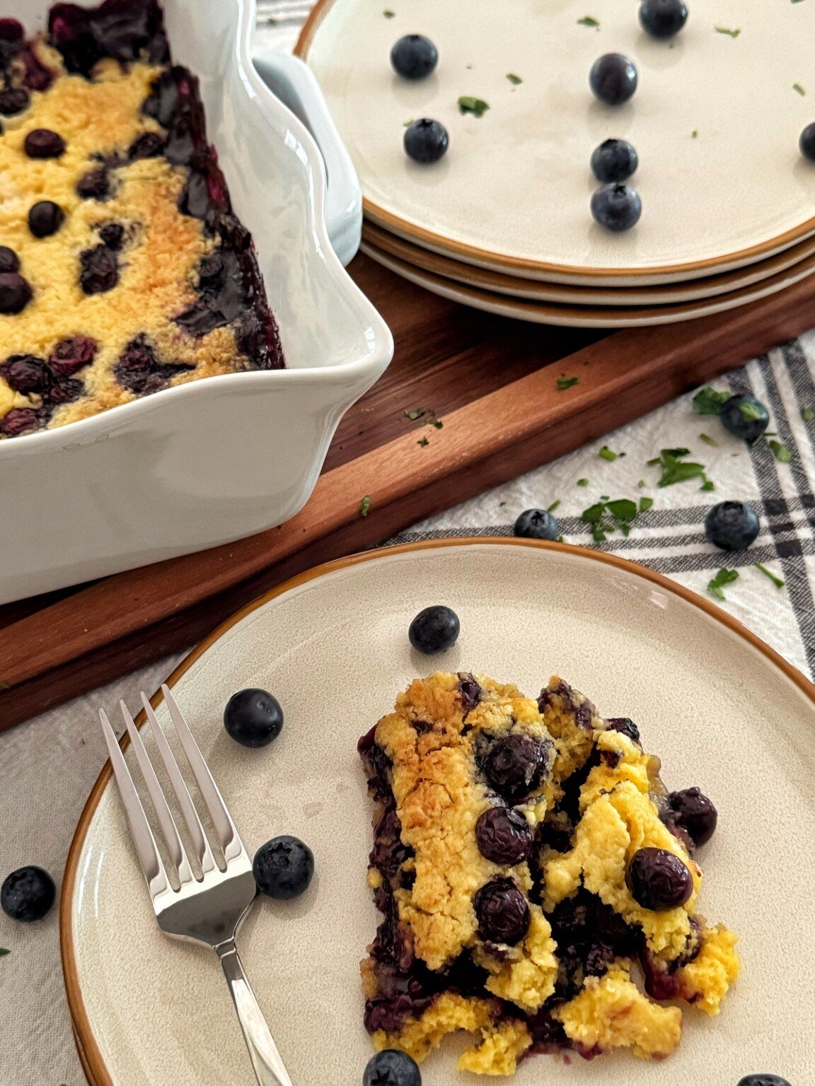 A slice of blueberry dump cake recipe on a plate with a fork, garnished with fresh blueberries. The baking dish with the remaining cobbler and stacked plates are in the background on a wooden board.