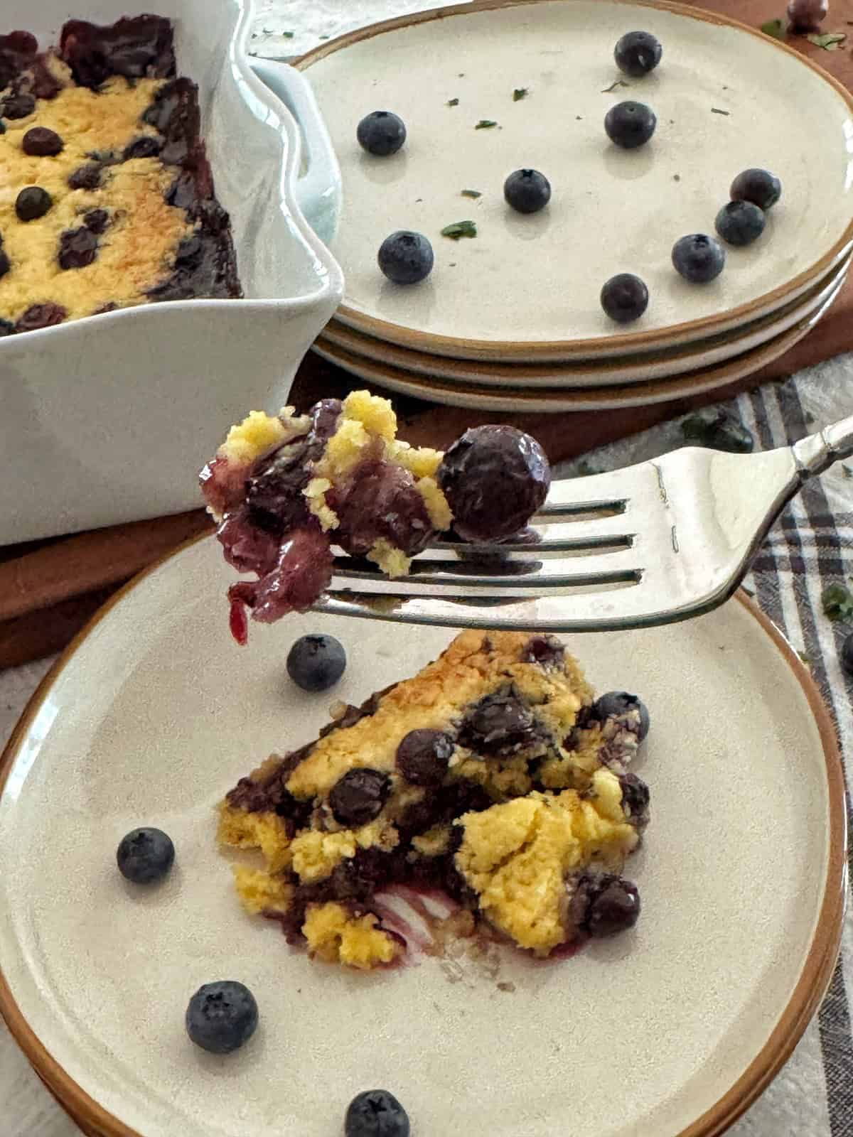 A close-up of a fork holding a bite of blueberry dump cake above a plate with a serving of cobbler and scattered fresh blueberries, with more plates, blueberries, and a baking dish in the background.